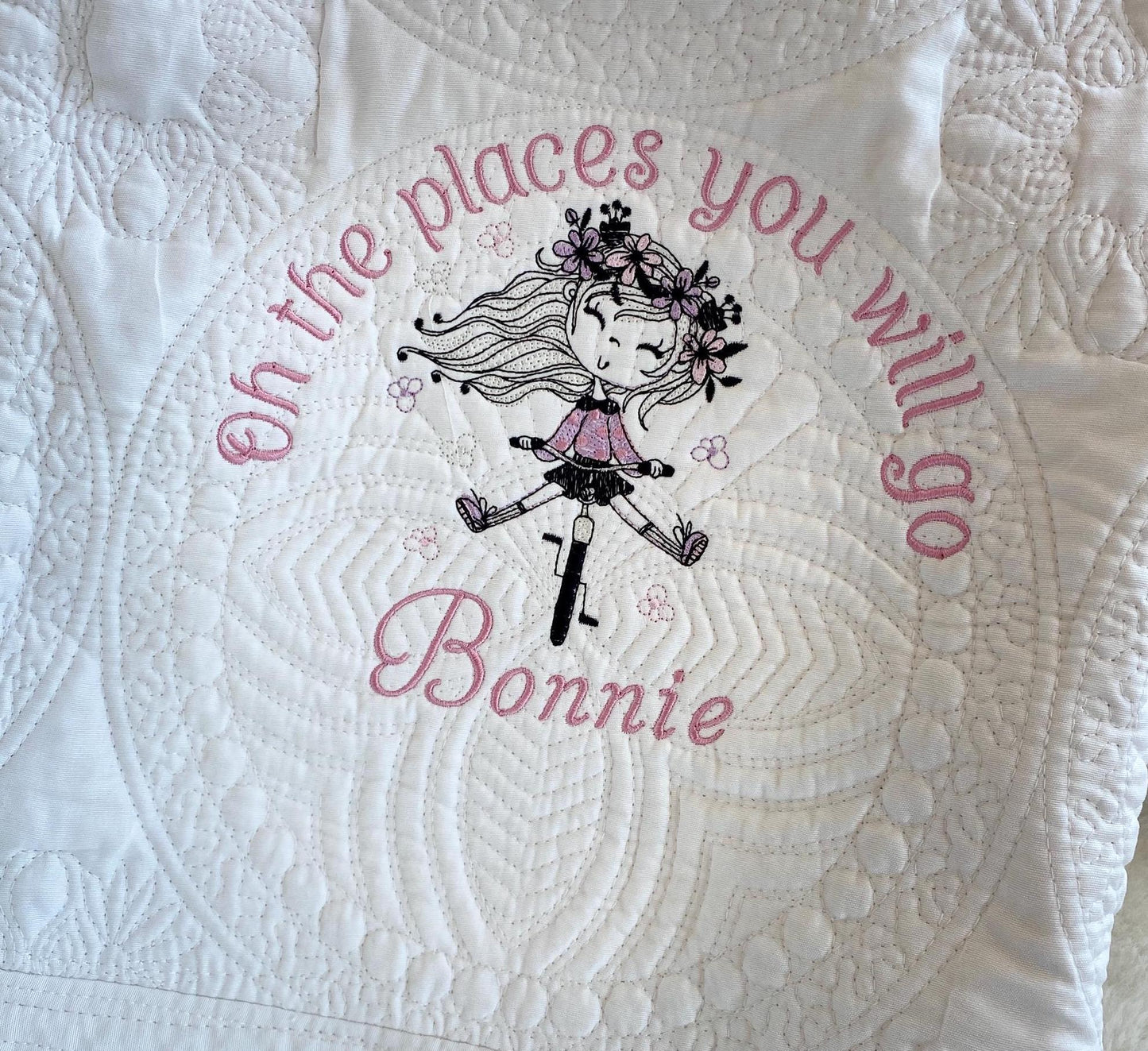 Close-up of a white Personalized Embroidered Girl on Bike Baby Quilt | Adventure Heirloom Keepsake | Custom Name Blanket featuring a stitched design of a girl on a bike with flowers and the phrase "Oh the places you will go" along with the custom name Bonnie.