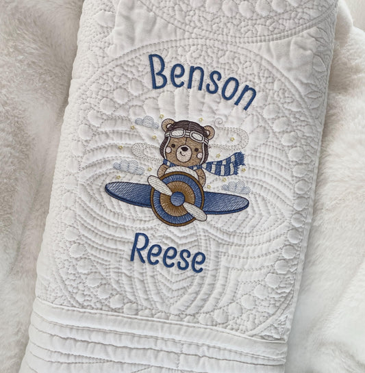 Close-up of a white quilt featuring detailed stitching and an embroidered bear pilot in blue and brown, personalized with the name "Benson Reese," from the Personalized Bear Pilot Airplane Baby Quilt | Custom Name Heirloom Blanket collection.