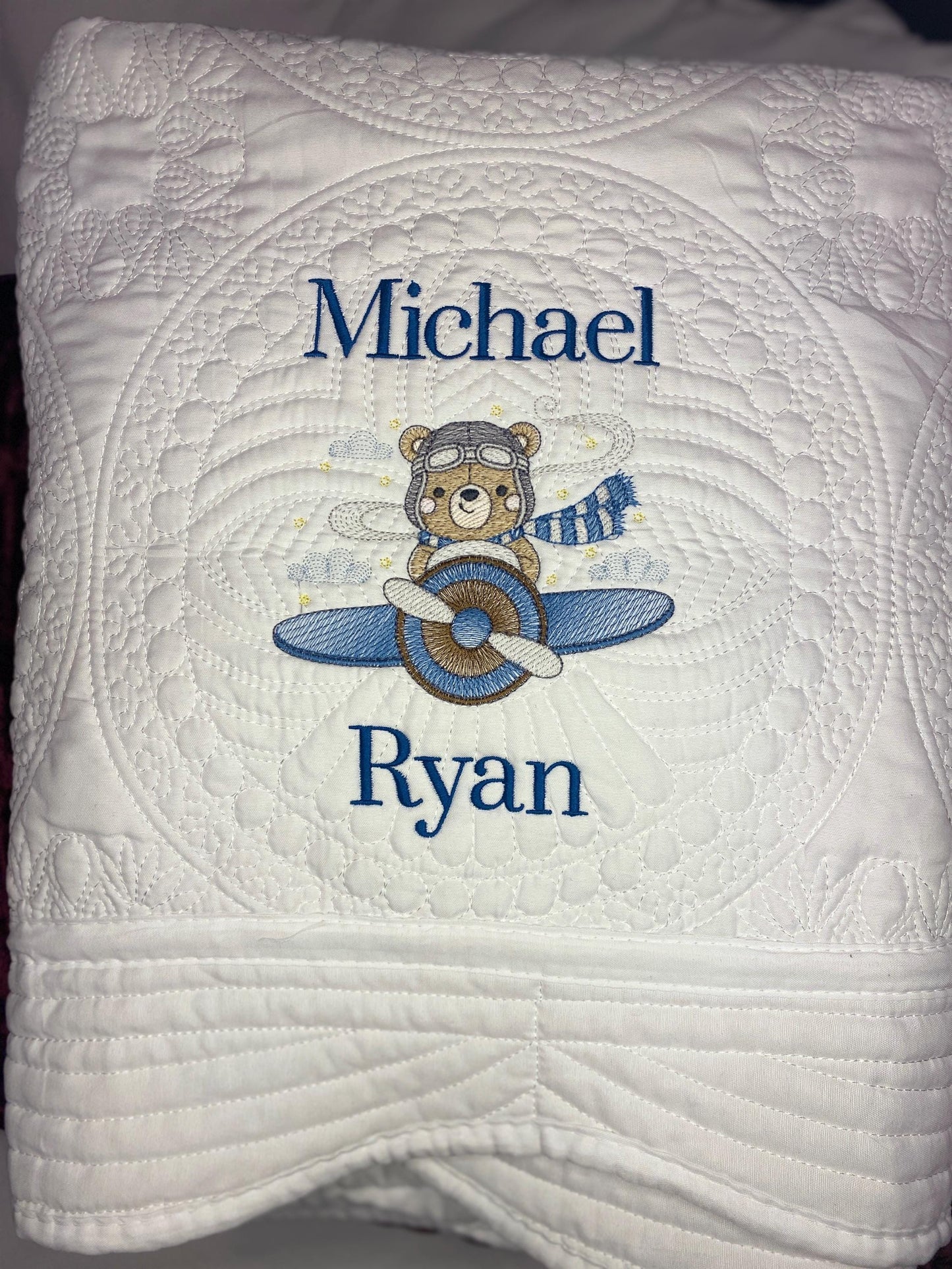 Personalized Bear Pilot Airplane Baby Quilt | Custom Name Heirloom Blanket with embroidered bear wearing pilot goggles and scarf flying a blue airplane, custom name "Michael Ryan" in blue thread on white quilted fabric.
