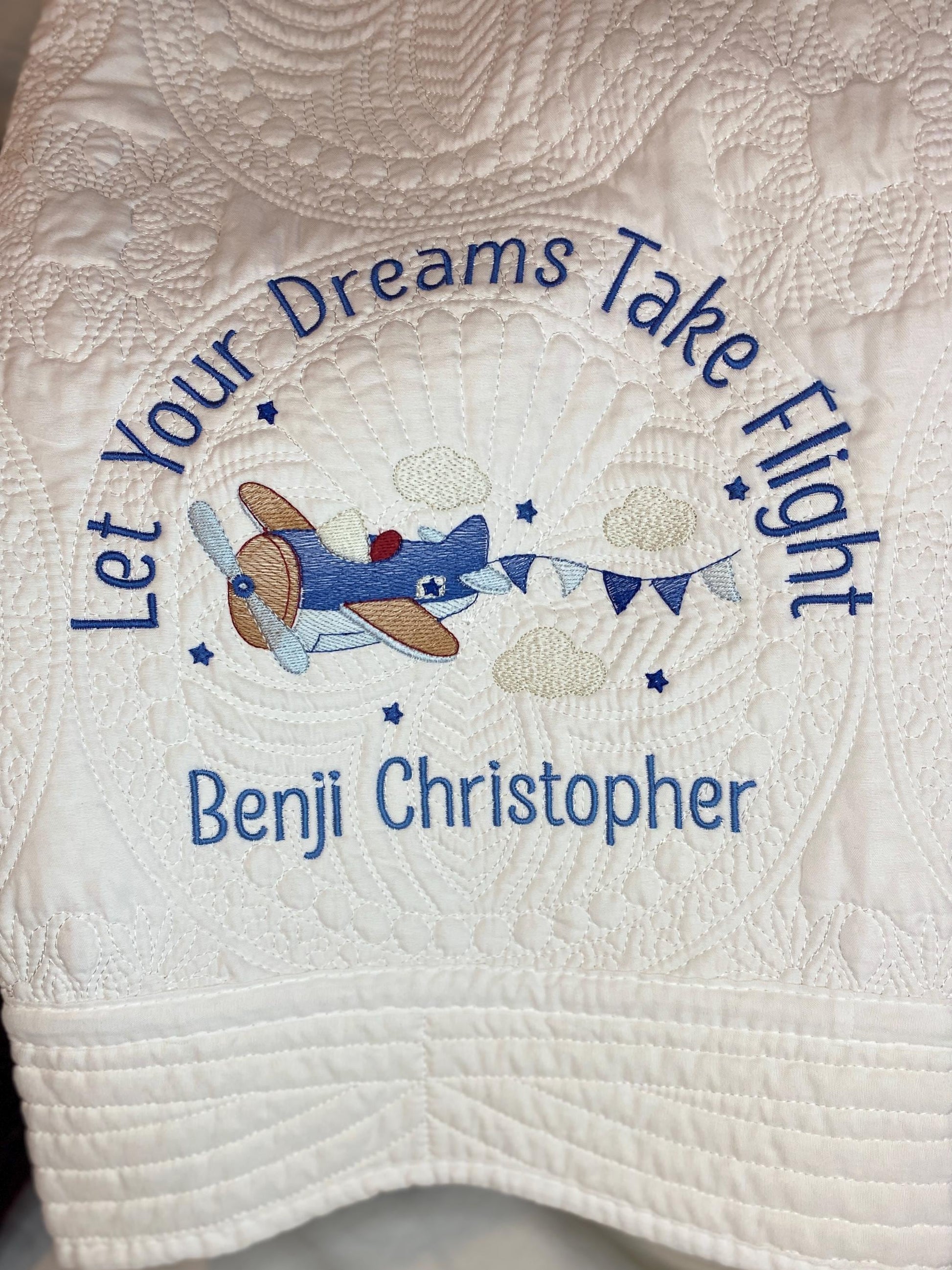 Personalized "Let Your Dreams Take Flight" Airplane Baby Quilt | Custom Name Blanket | Heirloom Keepsake featuring embroidered vintage airplane with clouds and custom name Benji Christopher on a white quilted background.
