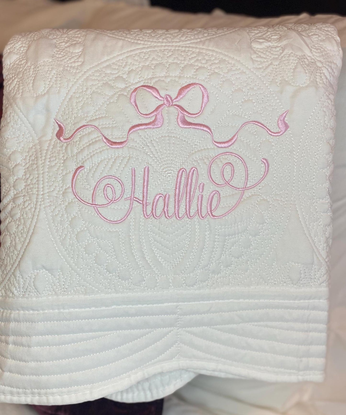 Personalized Pastel Pink Ribbon Bow Baby Quilt | Custom Name Heirloom Blanket for Girls with delicate pink embroidered ribbon bow and custom name "Hallie" on soft white fabric.