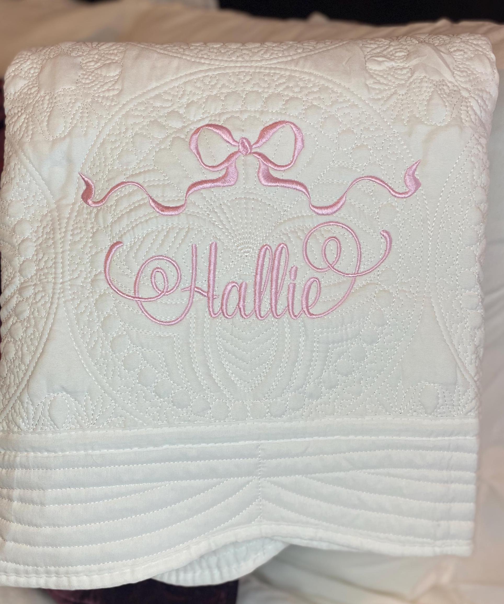 Personalized Pastel Pink Ribbon Bow Baby Quilt | Custom Name Heirloom Blanket for Girls with delicate pink embroidered ribbon bow and custom name "Hallie" on soft white fabric.