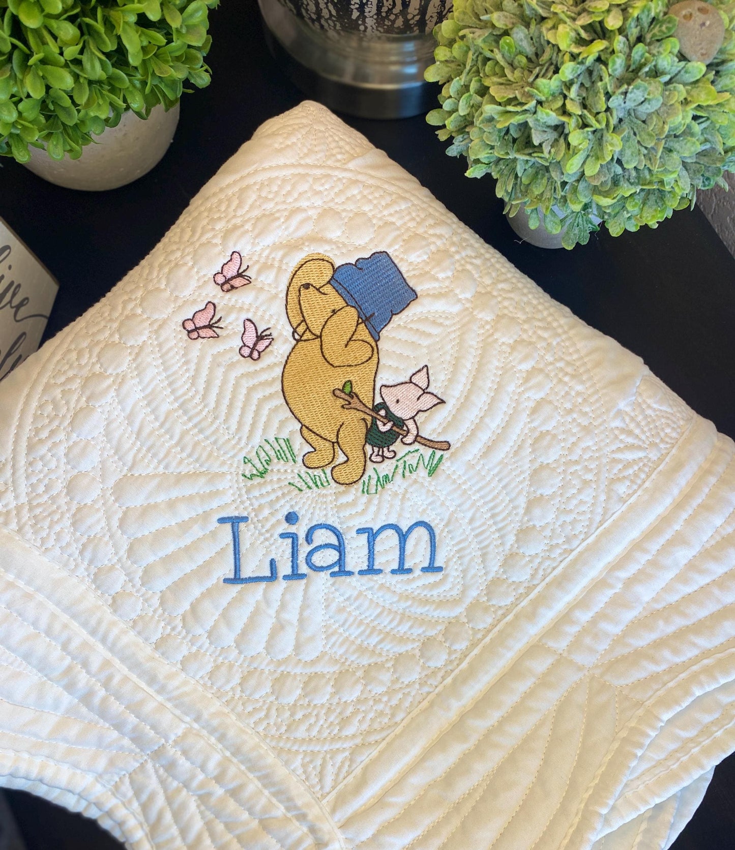 Close-up of the Personalized Storybook Bear & Friend with Bees Baby Quilt | Custom Name Heirloom Keepsake showcasing embroidered bear, cat, bees, butterflies, and the name Liam in blue stitching on a cream quilted fabric.