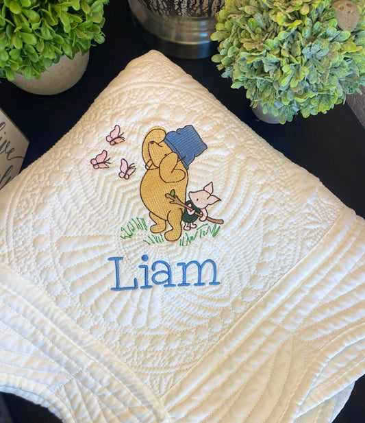 Close-up of the Personalized Storybook Bear & Friend with Bees Baby Quilt | Custom Name Heirloom Keepsake showcasing embroidered bear, cat, bees, butterflies, and the name Liam in blue stitching on a cream quilted fabric.