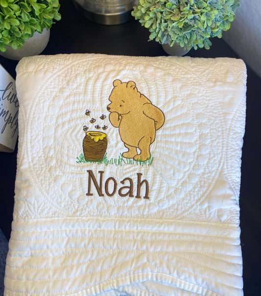 Personalized Storybook Bear & Honey Pot Baby Quilt | Custom Name Heirloom Keepsake featuring an embroidered bear with honey pot and custom name Noah on a soft white quilted blanket.