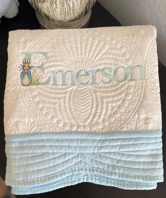 Personalized Classic Storybook Bunny Baby Quilt | Custom Name Heirloom Blanket featuring light blue embroidery of the name Emerson with a storybook bunny design on soft cream and blue fabric.