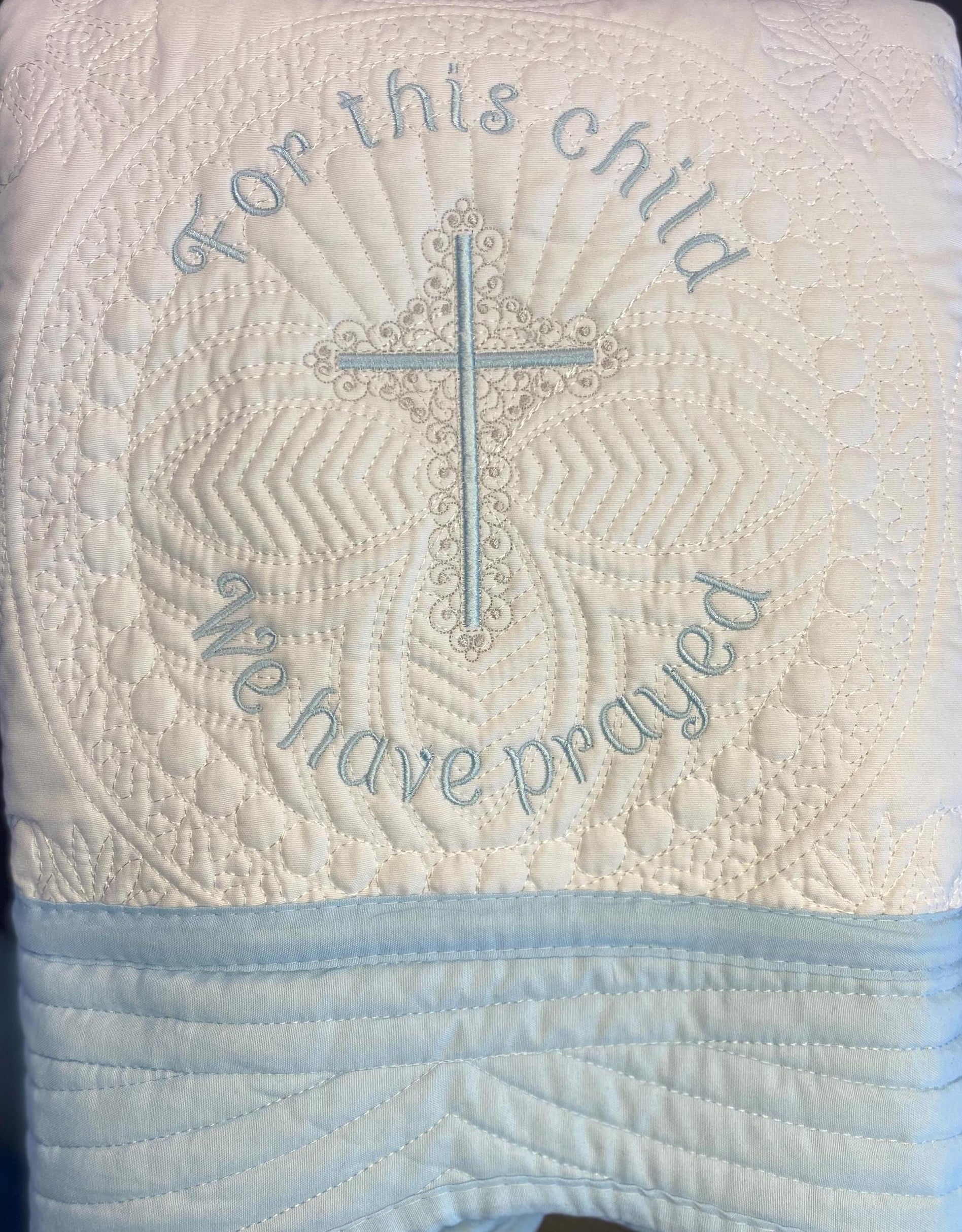 For This Child We Have Prayed" Cross Baby Quilt | Custom Embroidered Baptism Heirloom Blanket featuring intricate quilting with a blue embroidered cross and prayer message on a soft white fabric background.