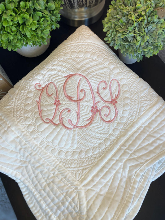 Personalized Vintage Satin Monogram Baby Quilt | Custom Embroidered Heirloom Blanket with delicate pink monogram embroidery on a white quilted satin fabric, styled alongside green plants.