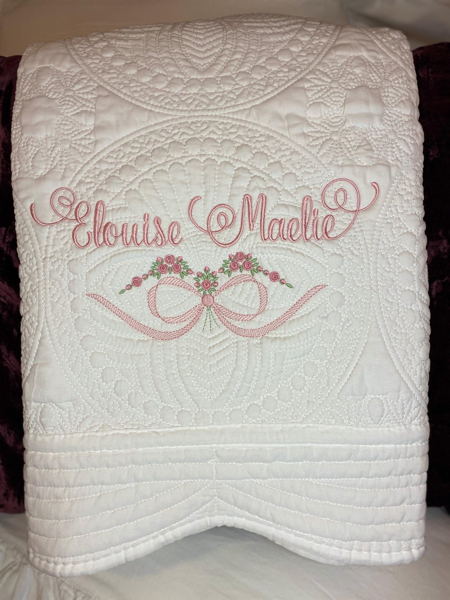 White quilt with intricate stitched patterns featuring custom pink embroidery of the names "Elouise Maelie" above a bow and rose design, showcasing the Personalized Roses & Bow Baby Quilt | Custom Embroidered Heirloom Blanket for Girls.