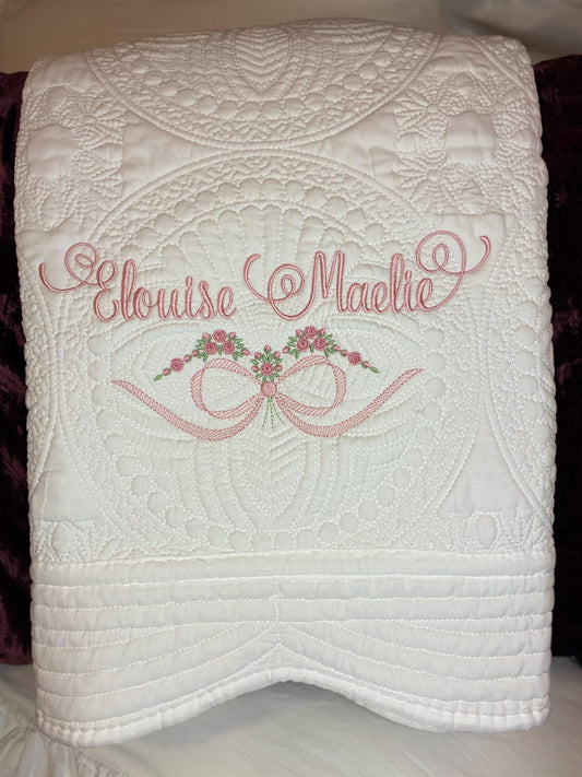 White quilt with intricate stitched patterns featuring custom pink embroidery of the names "Elouise Maelie" above a bow and rose design, showcasing the Personalized Roses & Bow Baby Quilt | Custom Embroidered Heirloom Blanket for Girls.