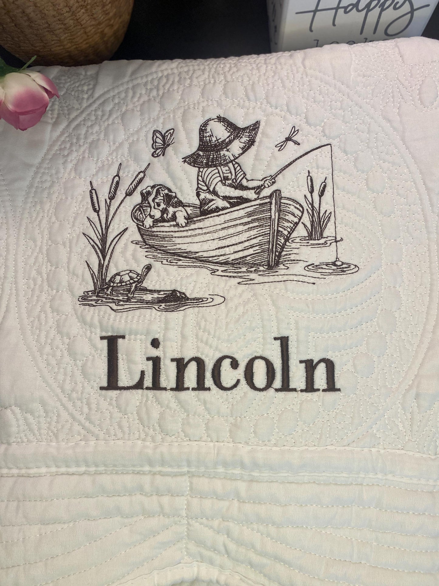 Embroidered scene of a little boy fishing from a boat with a dog, surrounded by cattails, dragonflies, and a turtle, personalized with the name Lincoln on the Personalized Little Fisherman Boy & Dog Baby Quilt | Custom Name Heirloom Blanket for Boys.