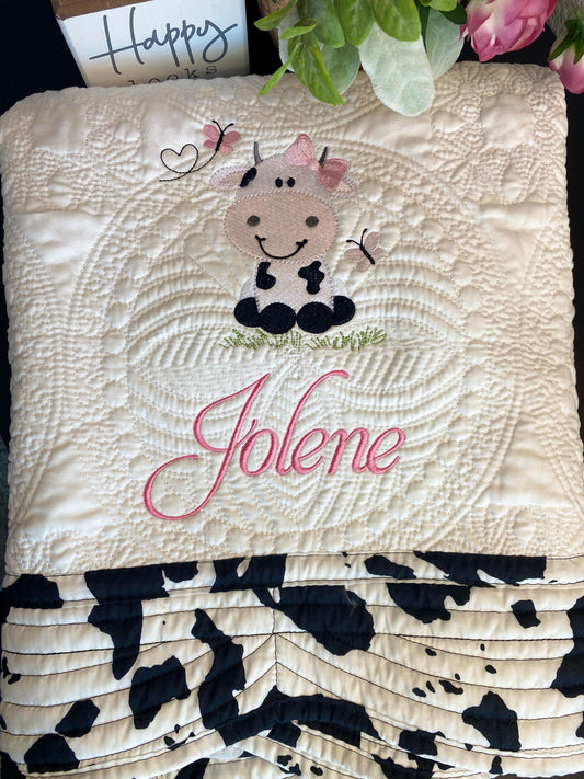 Personalized Embroidered Cow Baby Quilt | Custom Name Blanket | Heirloom Keepsake featuring a cute embroidered cow with a pink bow, butterflies, and the name Jolene stitched in pink on a soft quilted blanket with black and white cow print accents.
