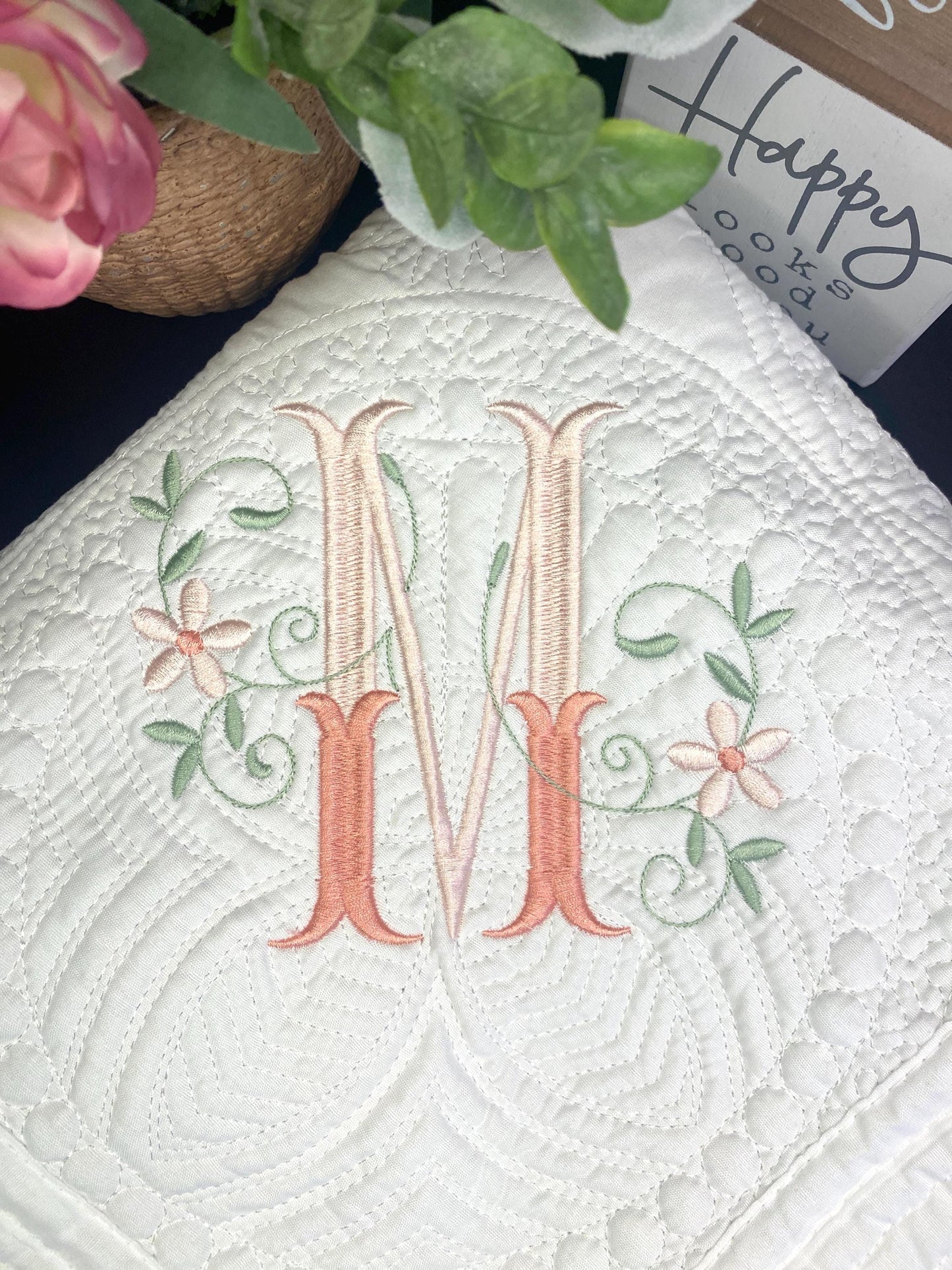 Close-up of a white quilt embroidered with an elegant floral interlocking monogram "M," showcasing delicate pink and green flower and vine details on the Personalized Elegant Floral Interlocking Monogram Baby Quilt | Custom Embroidered Heirloom Blanket.