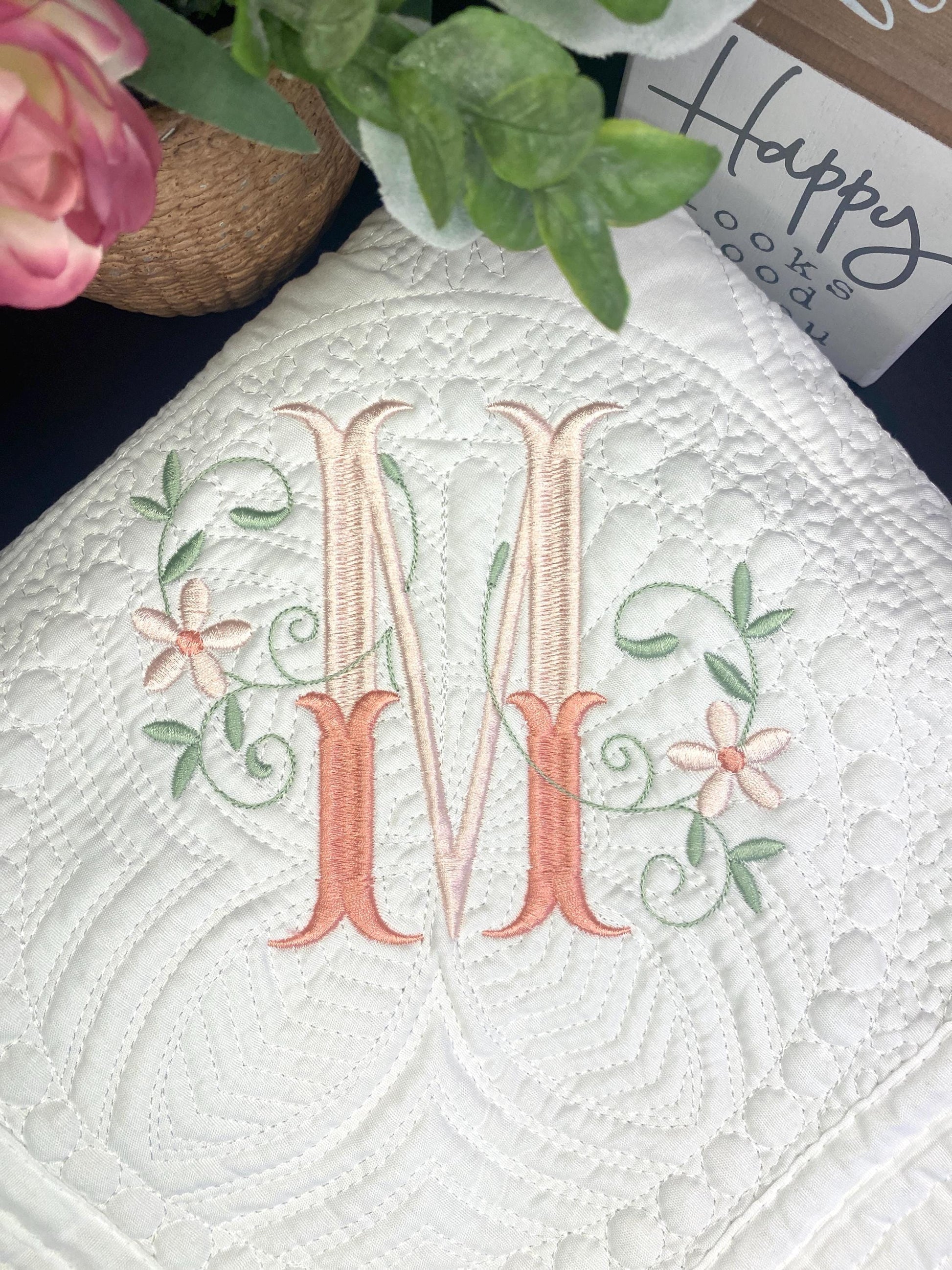 Close-up of a white quilt embroidered with an elegant floral interlocking monogram "M," showcasing delicate pink and green flower and vine details on the Personalized Elegant Floral Interlocking Monogram Baby Quilt | Custom Embroidered Heirloom Blanket.