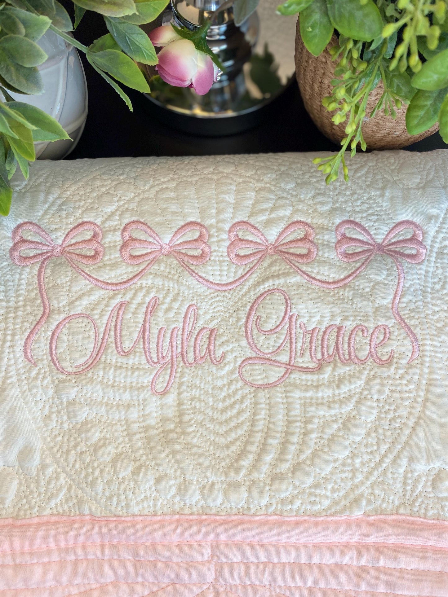 Personalized Pink Bow Baby Quilt | Custom Name Heirloom Blanket for Girls featuring delicate pink embroidered bows and the name Myla Grace on a soft quilted baby blanket surrounded by greenery.