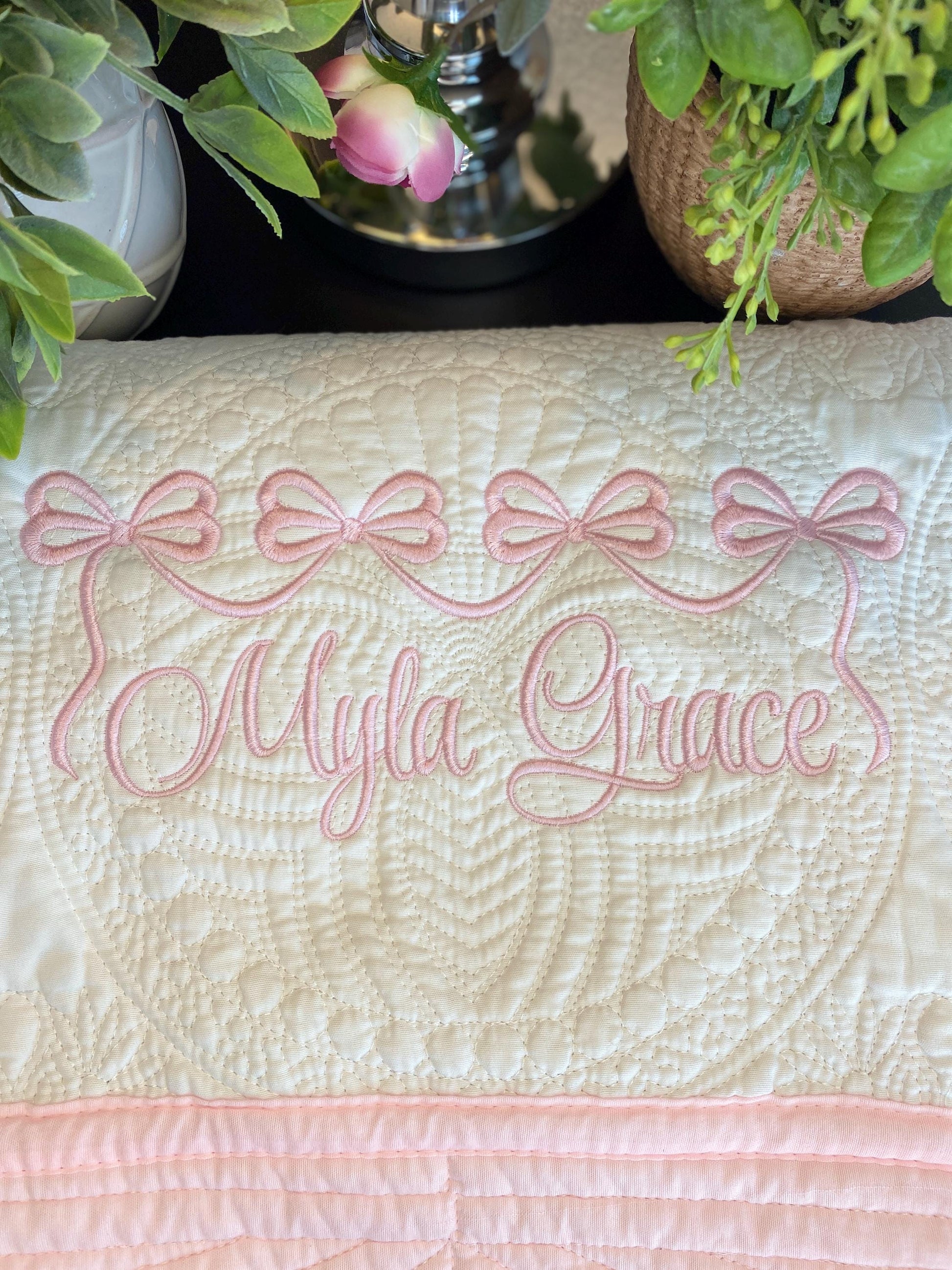 Personalized Pink Bow Baby Quilt | Custom Name Heirloom Blanket for Girls featuring delicate pink embroidered bows and the name Myla Grace on a soft quilted baby blanket surrounded by greenery.