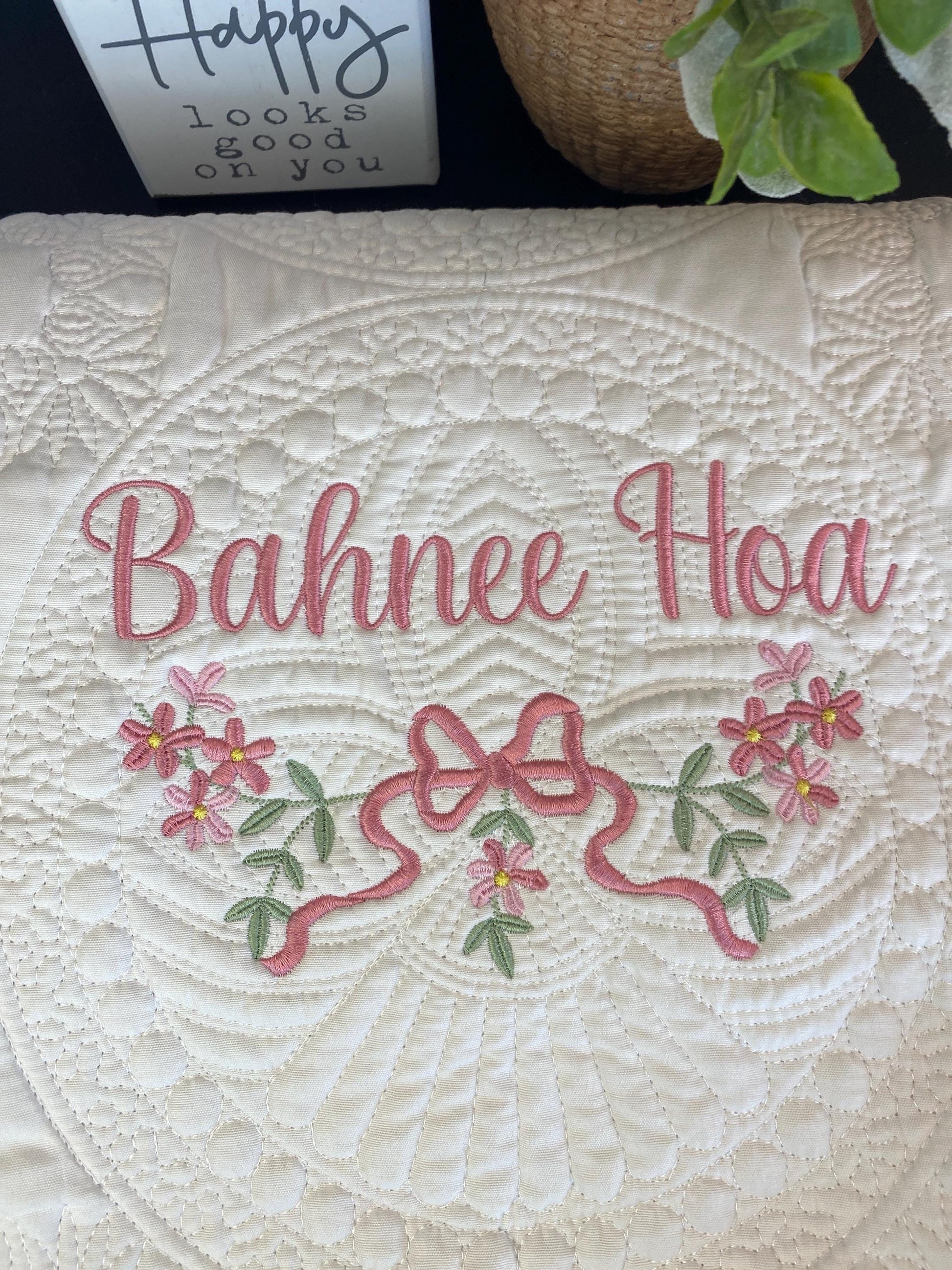 White quilt with intricate stitching and an embroidered pink bow and flower design surrounding the name "Bahnee Hoa," part of the Personalized Pink Flower & Bow Baby Quilt | Custom Name Heirloom Blanket for Girls.