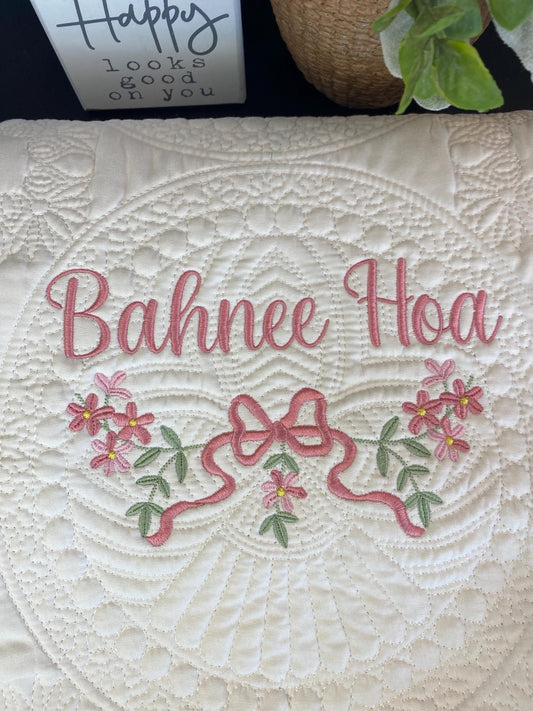White quilt with intricate stitching and an embroidered pink bow and flower design surrounding the name "Bahnee Hoa," part of the Personalized Pink Flower & Bow Baby Quilt | Custom Name Heirloom Blanket for Girls.