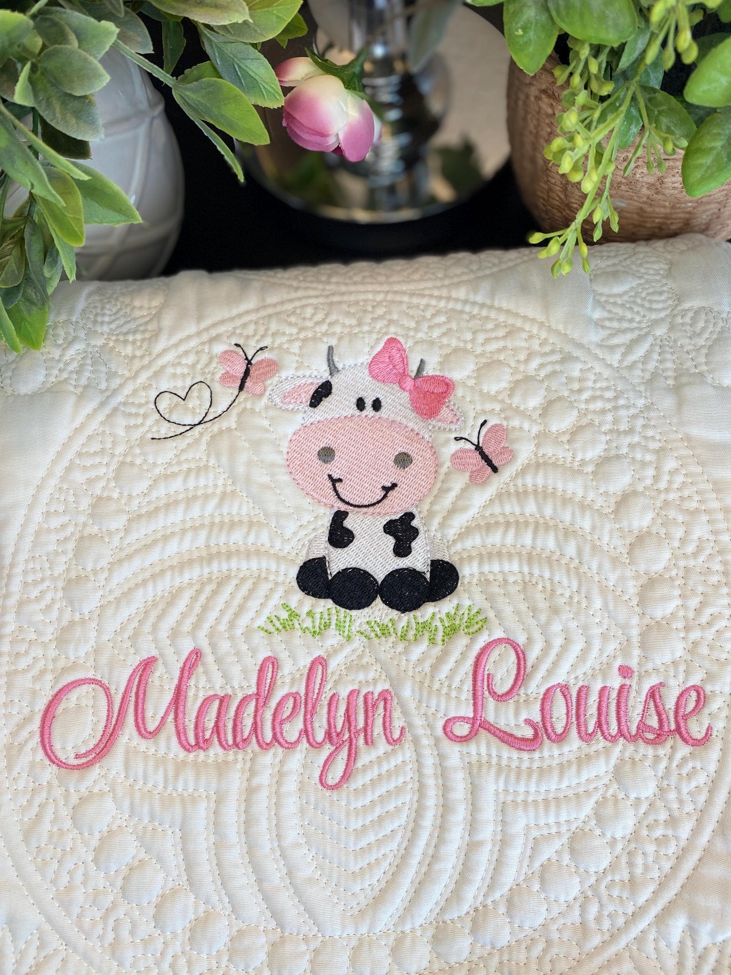 Close-up of a white Personalized Embroidered Cow Baby Quilt | Custom Name Blanket | Heirloom Keepsake featuring a cute embroidered cow with pink butterflies and the name "Madelyn Louise" stitched in pink thread.
