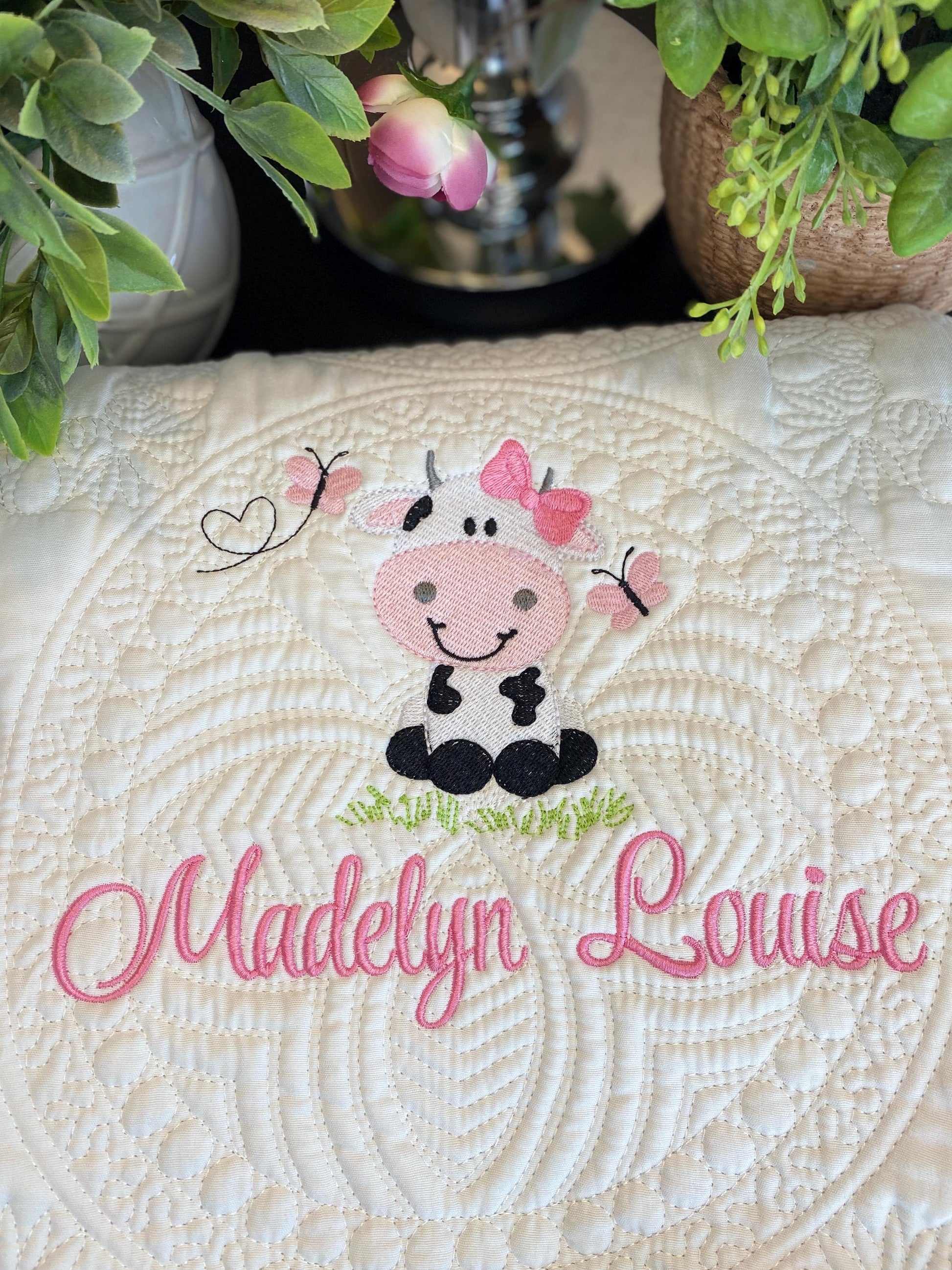 Close-up of a white Personalized Embroidered Cow Baby Quilt | Custom Name Blanket | Heirloom Keepsake featuring a cute embroidered cow with pink butterflies and the name "Madelyn Louise" stitched in pink thread.