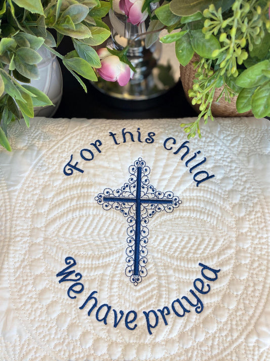 Detailed custom embroidered cross with the phrase "For this child we have prayed" on a cream quilt, showcasing the "For This Child We Have Prayed" Cross Baby Quilt | Custom Embroidered Baptism Heirloom Blanket surrounded by soft green and pink foliage.