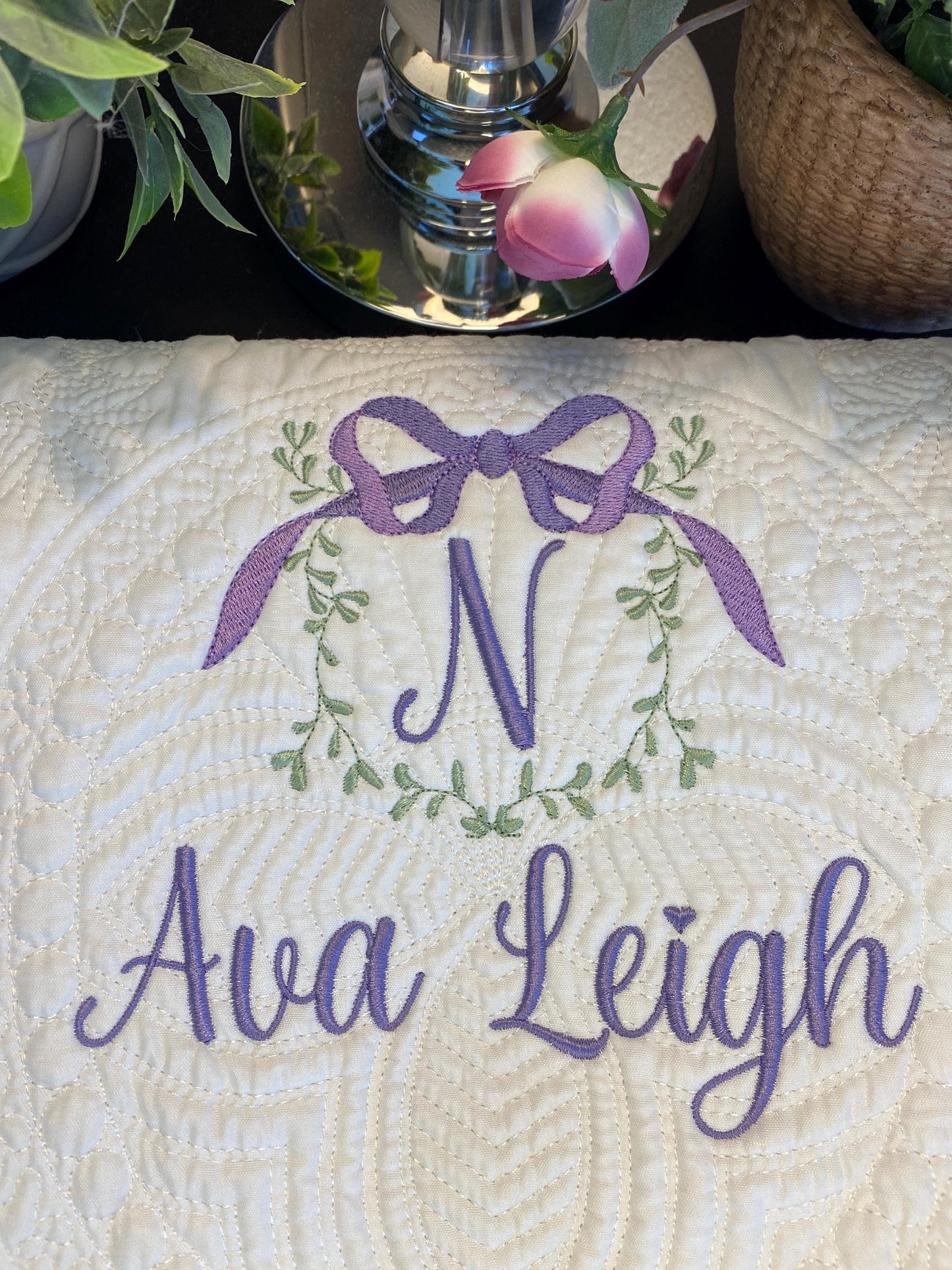 Personalized Vine & Bow Monogram Baby Quilt | Custom Embroidered Heirloom Blanket featuring a delicate purple bow and vine wreath monogram with custom name embroidery on a white quilted fabric.