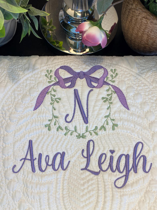 Personalized Vine & Bow Monogram Baby Quilt | Custom Embroidered Heirloom Blanket featuring a delicate purple bow and vine wreath monogram with custom name embroidery on a white quilted fabric.