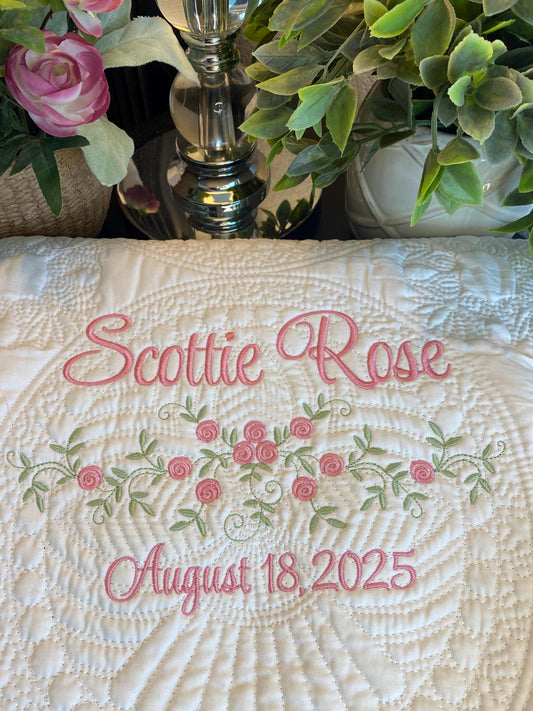 Personalized Delicate Floral Rose Swag Baby Quilt | Custom Embroidered Heirloom Blanket featuring pink embroidered name and date with intricate floral rose swag design on a white quilted background surrounded by green leaves and pink flowers.