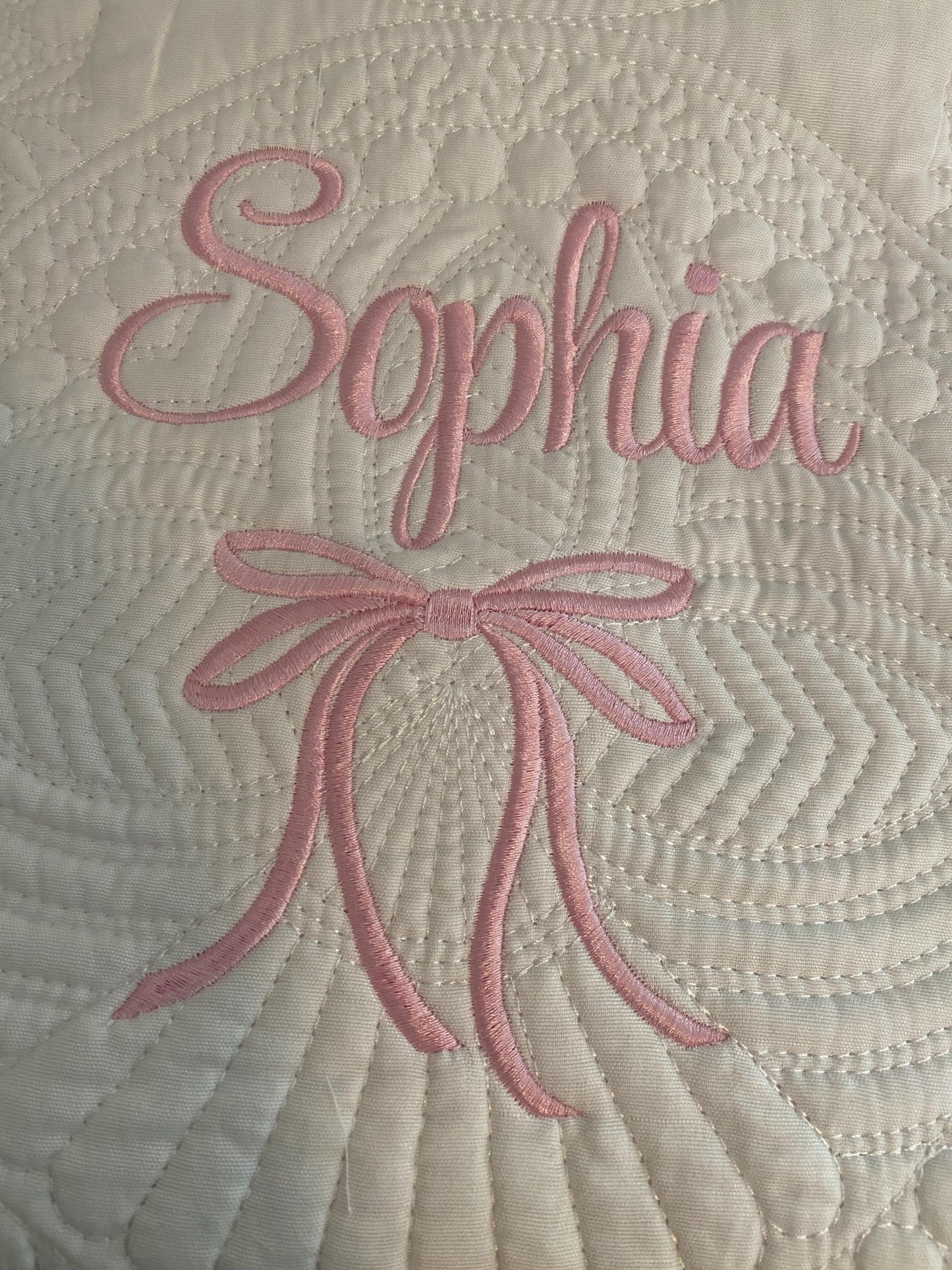 Close-up of a white quilt featuring intricate stitching with the name Sophia embroidered in pink above a matching embroidered bow, showcasing the Personalized Embroidered Bow Baby Quilt | Heirloom Keepsake | Custom Name Blanket.