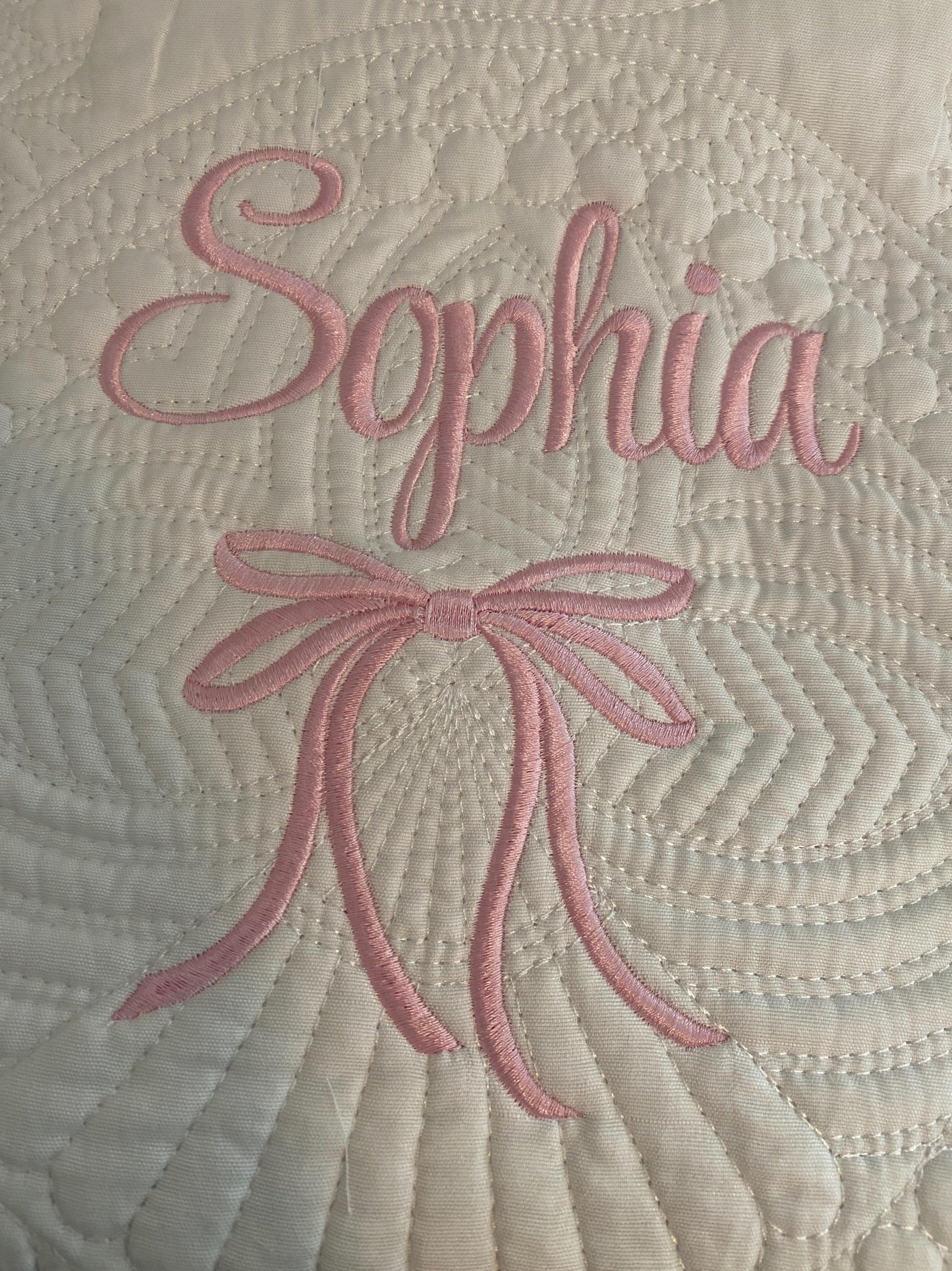 Close-up of a white quilt featuring intricate stitching with the name Sophia embroidered in pink above a matching embroidered bow, showcasing the Personalized Embroidered Bow Baby Quilt | Heirloom Keepsake | Custom Name Blanket.