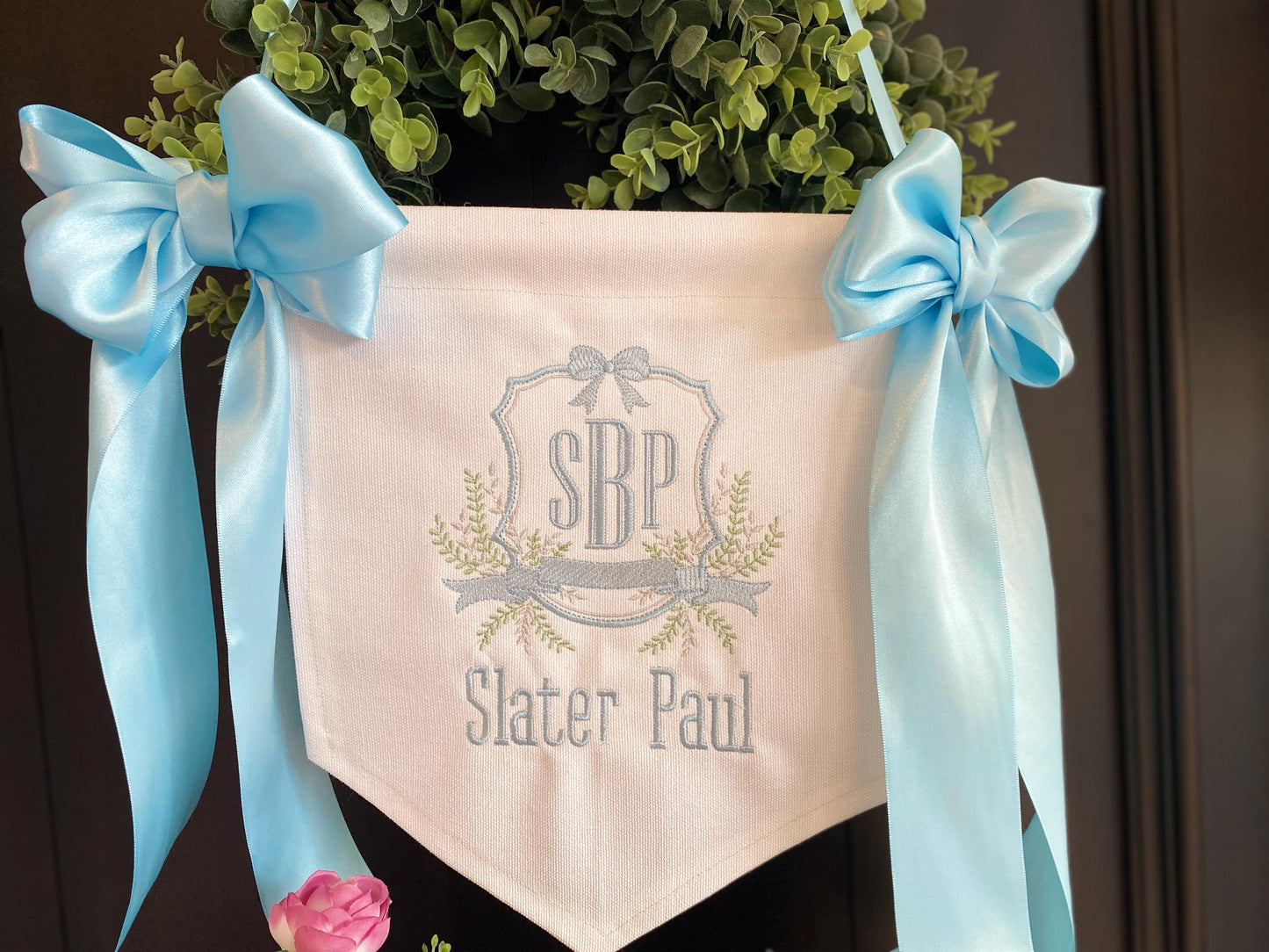 Personalized Embroidered Baby Birth Announcement Banner | Hospital Door & Bassinet Sign with light blue satin ribbons, custom monogram, and name Slater Paul on white fabric hanging against greenery.