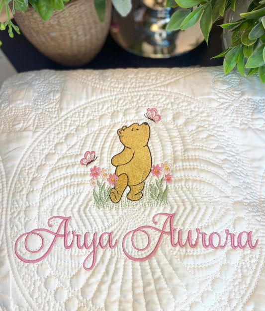 Embroidered bear with pink flowers and butterflies on a cream quilt featuring custom name embroidery, showcasing the Personalized Storybook Bear Pink Flowers & Butterflies Baby Quilt | Custom Name Heirloom Keepsake.