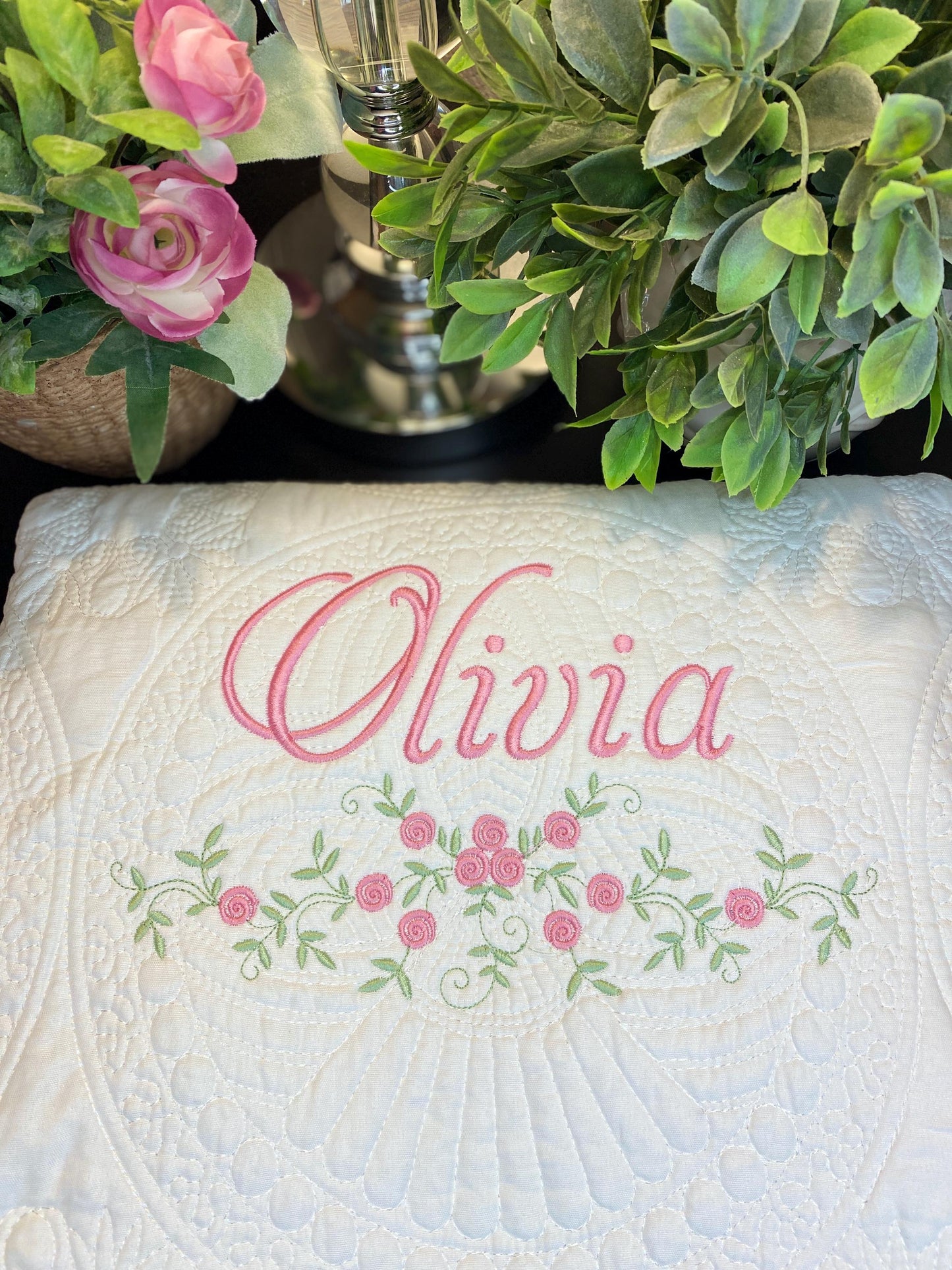 White quilt with custom embroidered name Olivia in pink script above a delicate floral rose swag design, surrounded by greenery, showcasing the Personalized Delicate Floral Rose Swag Baby Quilt | Custom Embroidered Heirloom Blanket.