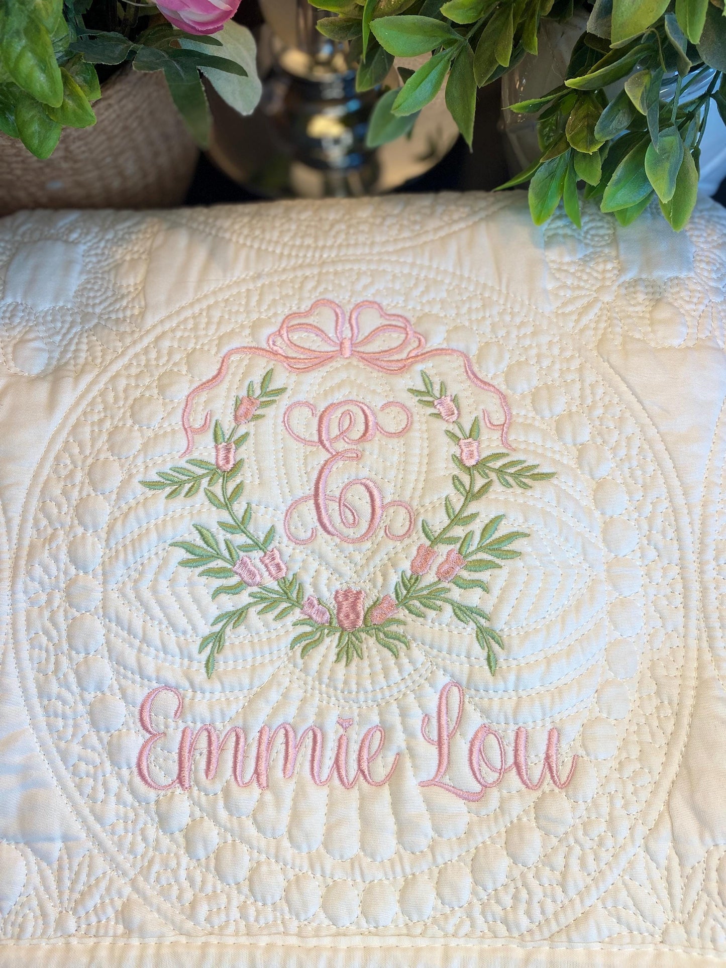 Close-up of a personalized bow and pink floral wreath monogram baby quilt featuring delicate embroidery with the letter E and the name Emmie Lou on a cream background surrounded by greenery, showcasing the "Personalized Bow & Pink Floral Wreath Monogram Baby Quilt | Custom Embroidered Heirloom Blanket" craftsmanship.