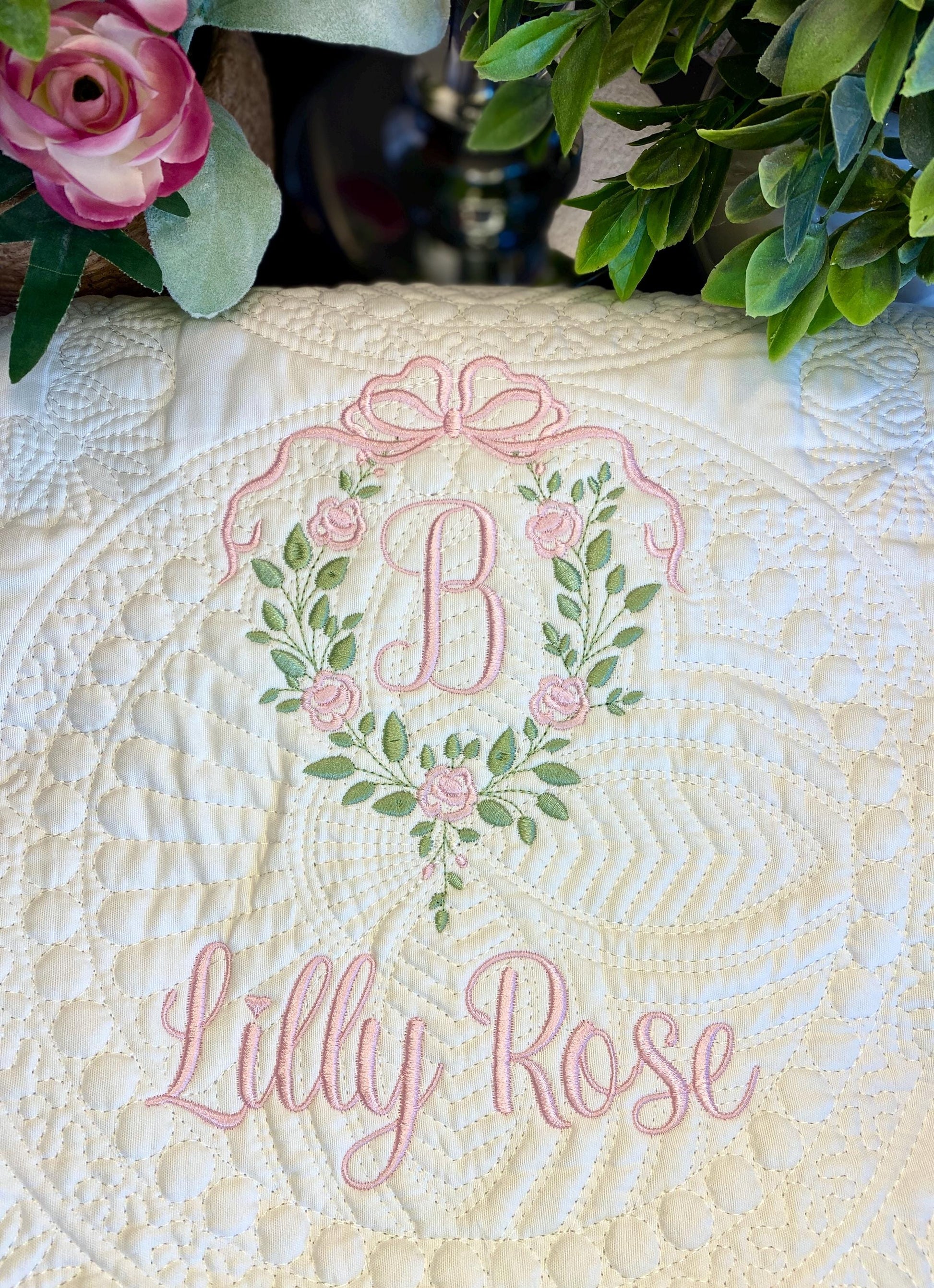 Personalized Rose Vine Bow Crest Monogram Baby Quilt | Custom Embroidered Heirloom Blanket featuring a pink embroidered monogram "B" surrounded by a rose vine and bow, with the name Lilly Rose stitched below on a cream quilted fabric.