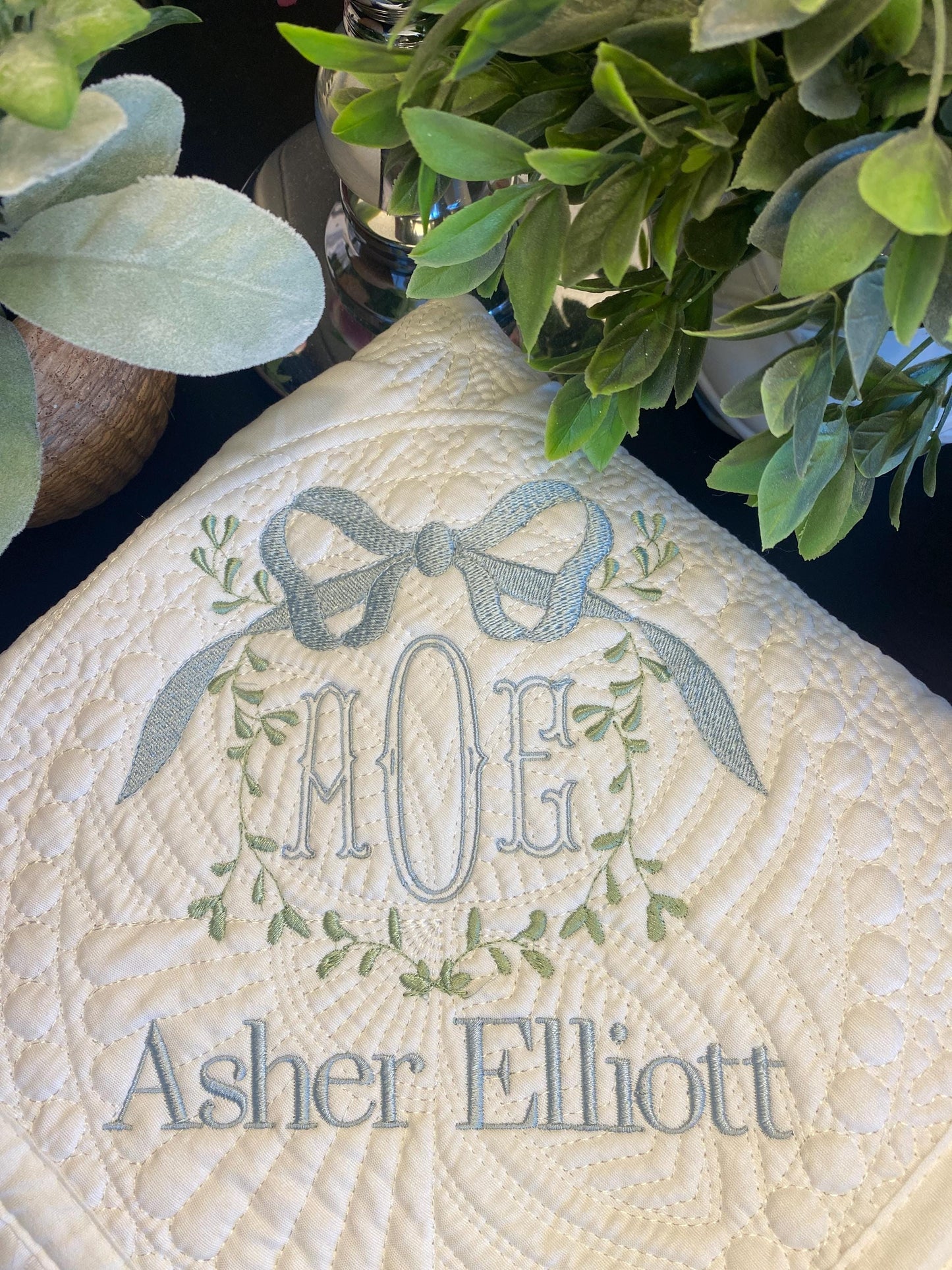 Close-up of an embroidered monogram with initials AOG topped by a vine and bow design and the name Asher Elliott on a white quilted surface, Personalized Vine Bow Crest Monogram Baby Quilt | Custom Embroidered Heirloom Keepsake.