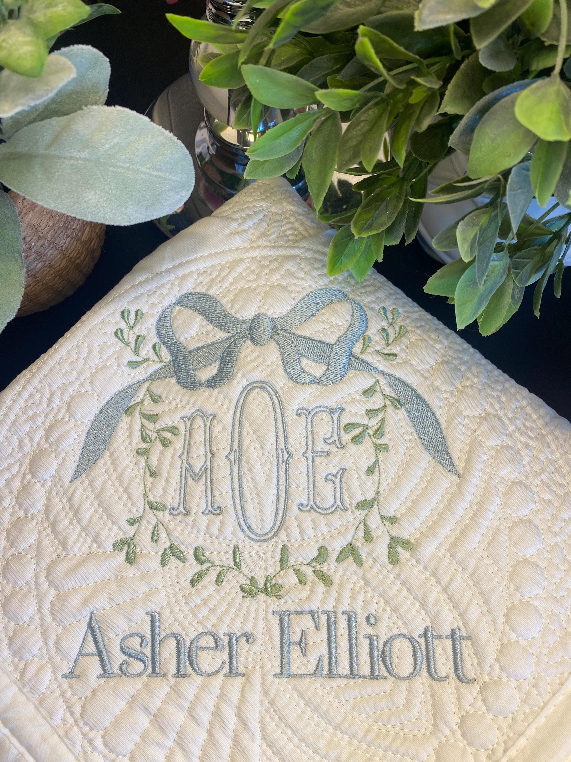 Close-up of an embroidered monogram with initials AOG topped by a vine and bow design and the name Asher Elliott on a white quilted surface, Personalized Vine Bow Crest Monogram Baby Quilt | Custom Embroidered Heirloom Keepsake.