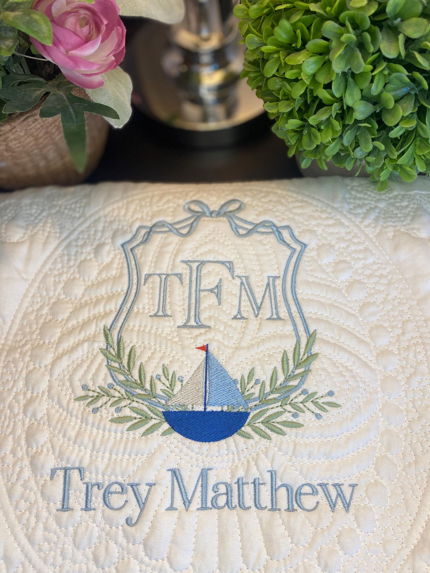 Close-up of a Personalized Sailboat Crest Monogram Baby Quilt | Custom Embroidered Heirloom Blanket for Boys featuring a blue sailboat, green laurel leaves, and custom embroidered name Trey Matthew on a cream quilted fabric.