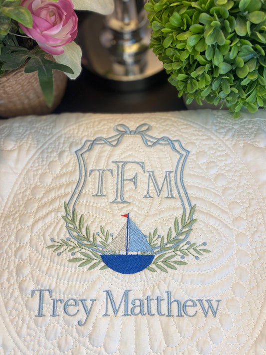 Close-up of a Personalized Sailboat Crest Monogram Baby Quilt | Custom Embroidered Heirloom Blanket for Boys featuring a blue sailboat, green laurel leaves, and custom embroidered name Trey Matthew on a cream quilted fabric.
