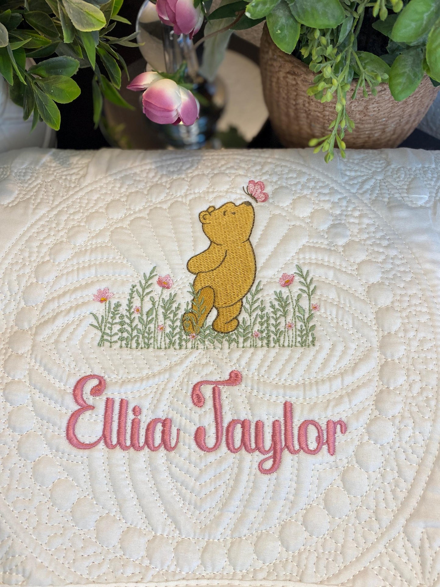 Personalized Classic Pooh Bear Meadow & Butterfly Baby Quilt | Custom Name Heirloom Blanket featuring an embroidered Pooh bear with pink butterfly and flowers above the custom name Ellia Taylor in pink script on a soft quilted white background.