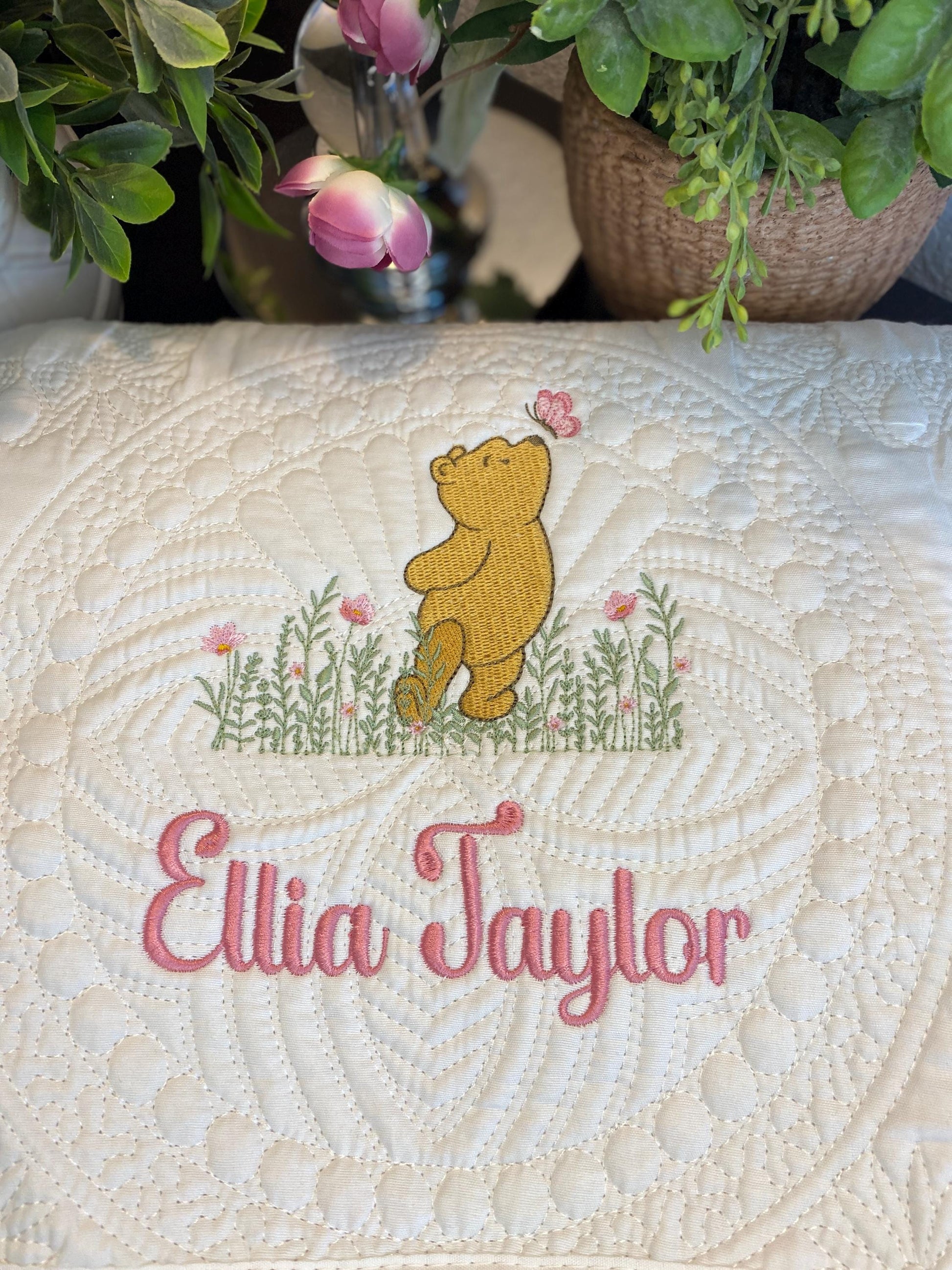 Personalized Classic Pooh Bear Meadow & Butterfly Baby Quilt | Custom Name Heirloom Blanket featuring an embroidered Pooh bear with pink butterfly and flowers above the custom name Ellia Taylor in pink script on a soft quilted white background.