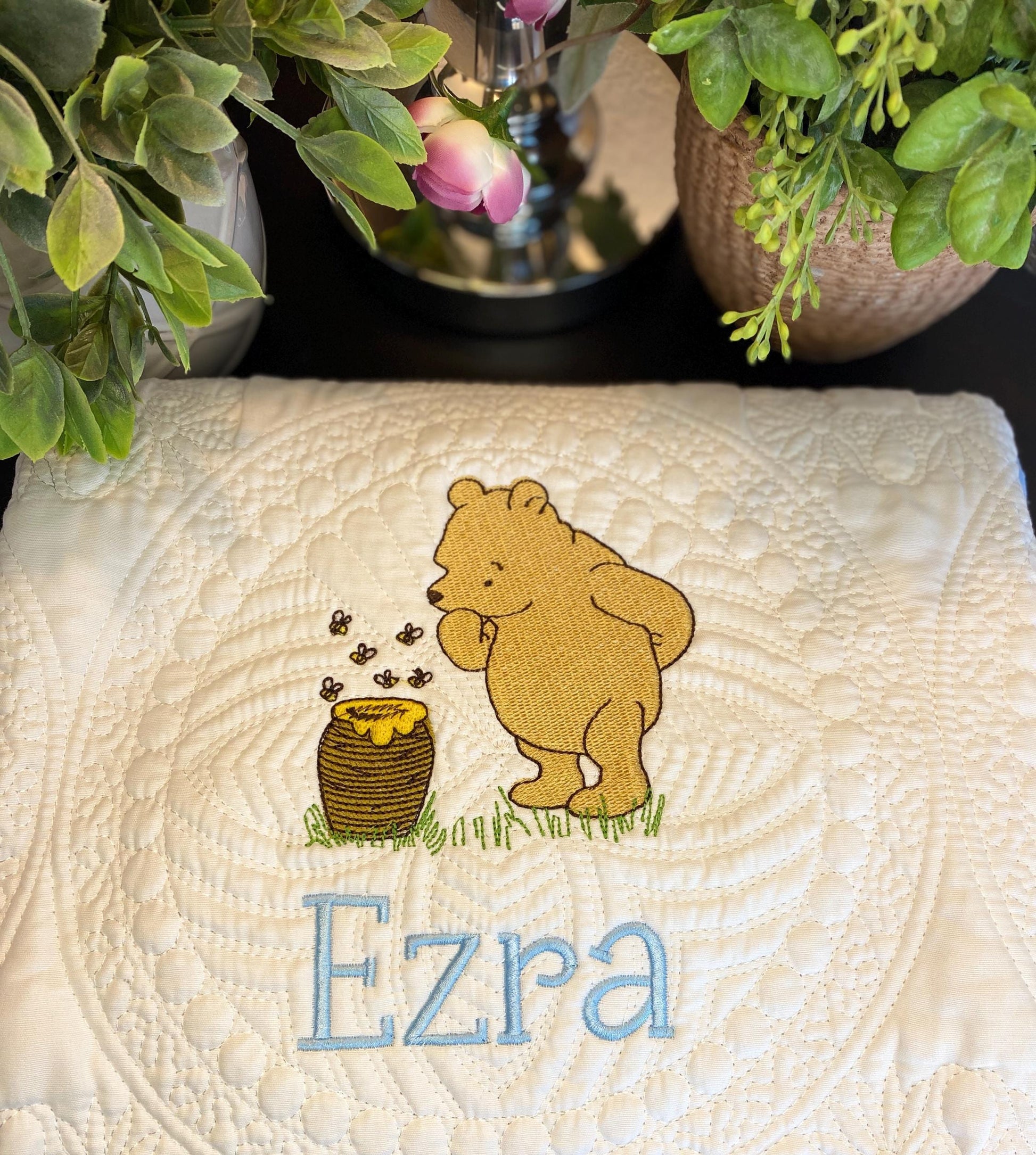 Personalized Storybook Bear & Honey Pot Baby Quilt | Custom Name Heirloom Keepsake featuring an embroidered bear and honey pot design with the name Ezra in blue thread on a quilted white blanket.