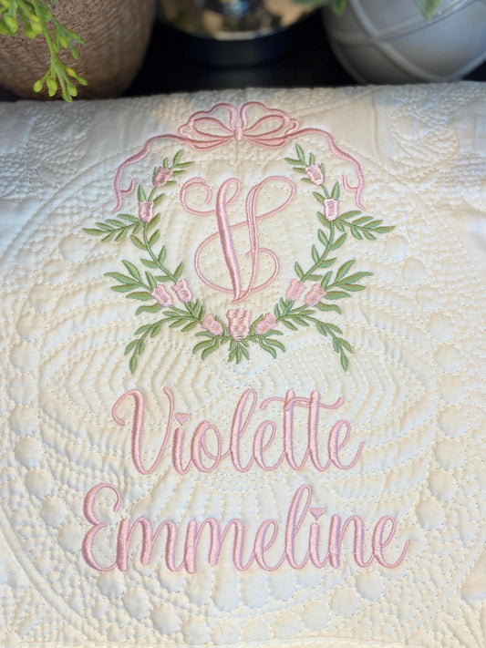 Personalized Bow & Pink Floral Wreath Monogram Baby Quilt | Custom Embroidered Heirloom Blanket with a delicate pink bow and floral wreath embroidery surrounding a monogram, featuring the name Violette Emmeline on a soft quilted fabric.
