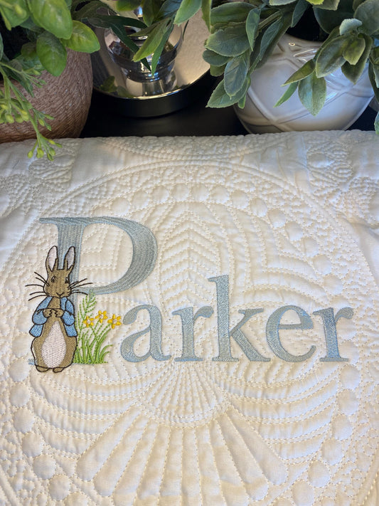 Close-up of a white Personalized Classic Storybook Bunny Baby Quilt | Custom Name Heirloom Blanket featuring embroidered gentle blue lettering and a bunny holding flowers, surrounded by quilted patterns and greenery.