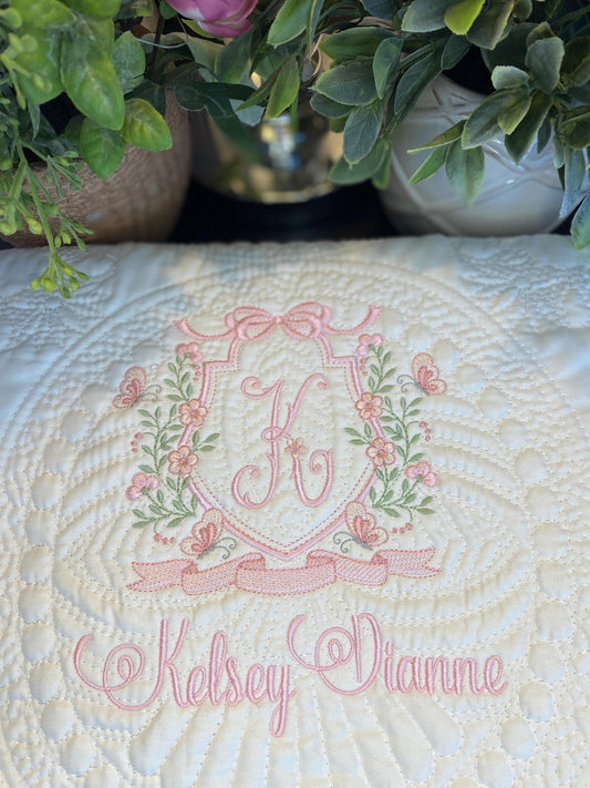 Personalized Pink Floral Vine Bow Crest Monogram Baby Quilt | Custom Embroidered Heirloom Blanket featuring delicate pink embroidery of a vine, bow, butterflies, and a scripted monogram with the name Kelsey Dianne on a soft white quilt.