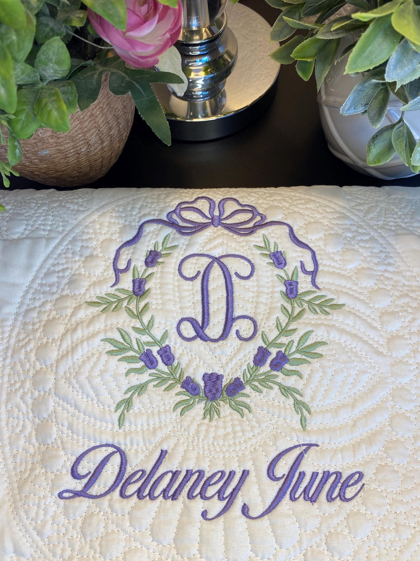 Personalized Bow & Pink Floral Wreath Monogram Baby Quilt | Custom Embroidered Heirloom Blanket featuring a delicate purple embroidered wreath and monogram D with the name Delaney June on a quilted cream background.