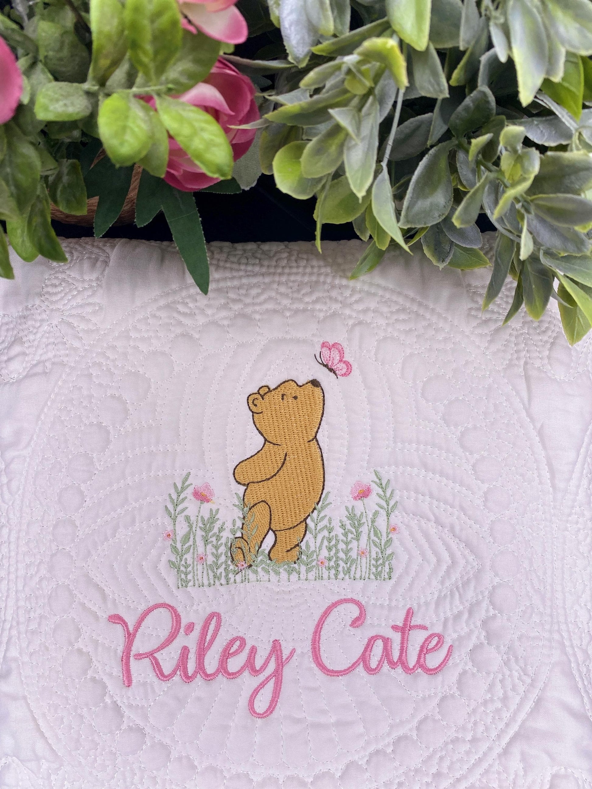 Personalized Classic Pooh Bear Meadow & Butterfly Baby Quilt | Custom Name Heirloom Blanket featuring embroidery of a bear reaching for a butterfly over flowers with custom name Riley Cate in pink script.