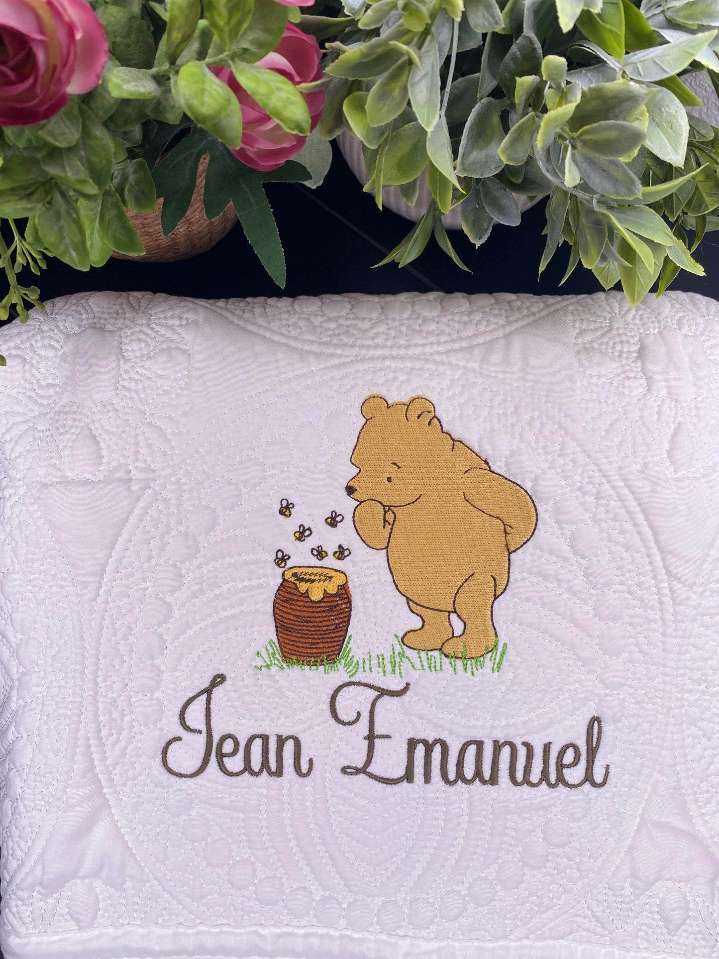 Personalized Storybook Bear & Honey Pot Baby Quilt | Custom Name Heirloom Keepsake featuring an embroidered storybook bear with honey pot and custom name on a white quilted background surrounded by greenery and flowers.