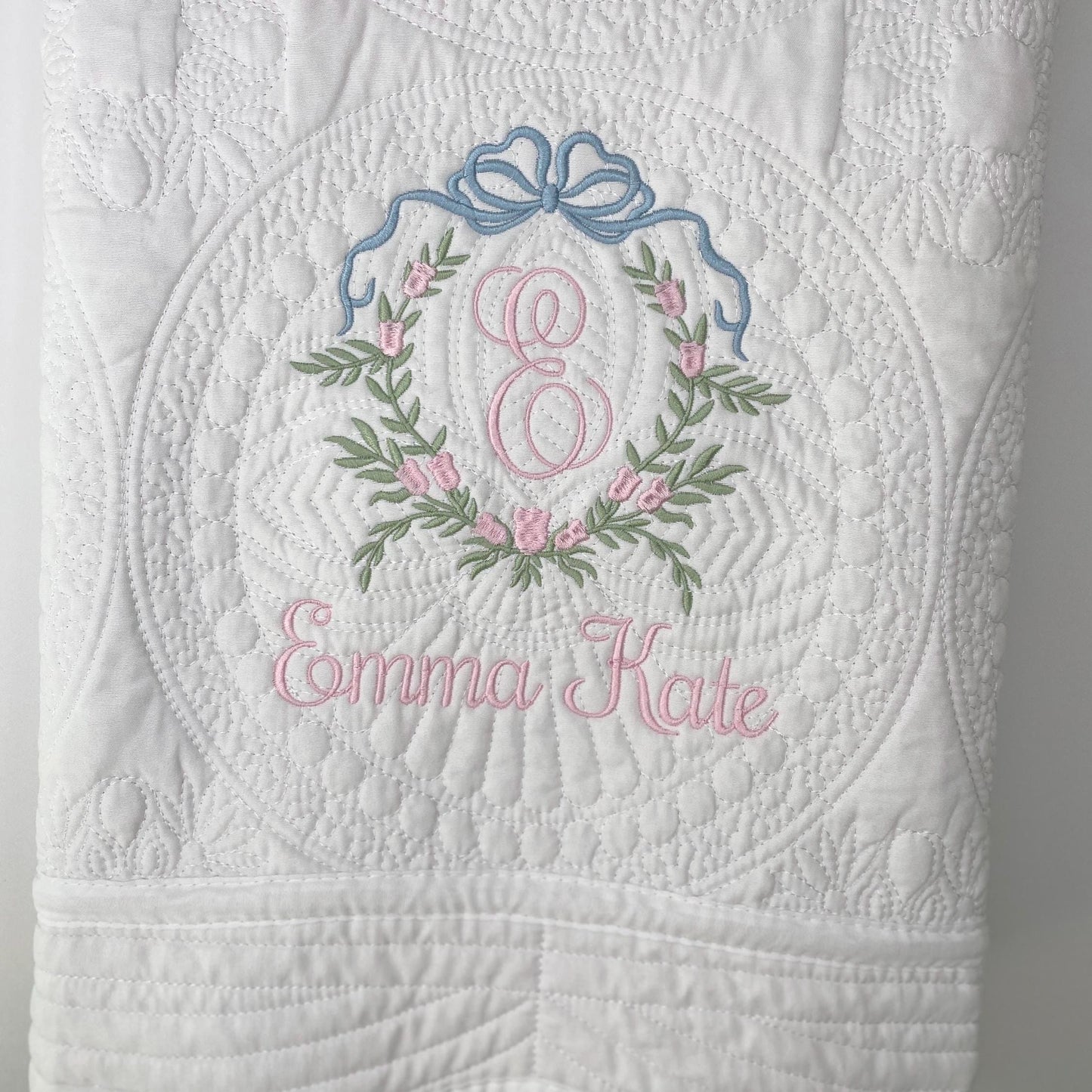 White quilt with detailed floral and bow embroidery in pink and blue, featuring a pink monogram "E" and the name Emma Kate, showcasing the Personalized Bow & Pink Floral Wreath Monogram Baby Quilt | Custom Embroidered Heirloom Blanket design.