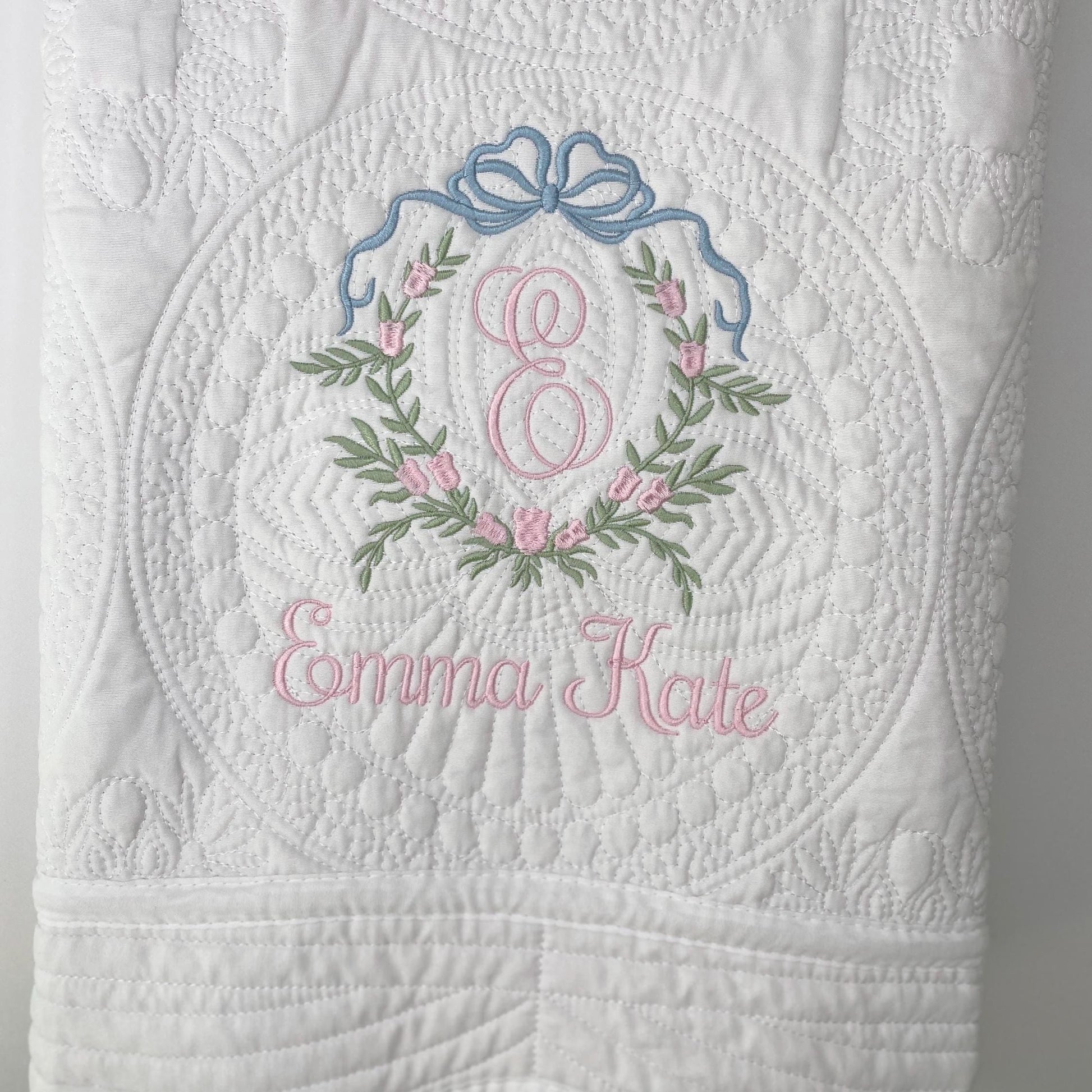 White quilt with detailed floral and bow embroidery in pink and blue, featuring a pink monogram "E" and the name Emma Kate, showcasing the Personalized Bow & Pink Floral Wreath Monogram Baby Quilt | Custom Embroidered Heirloom Blanket design.